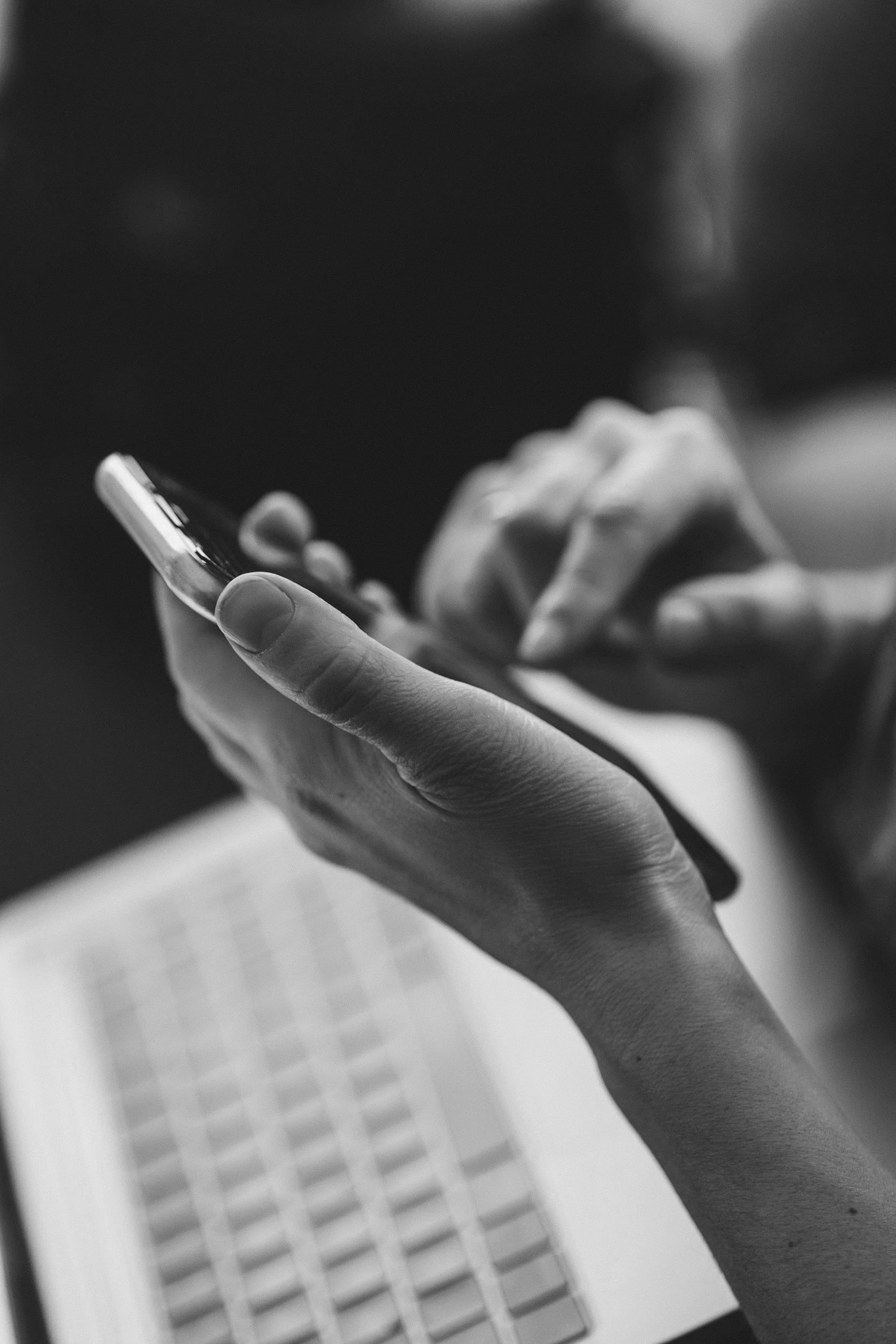 Black and white photo of hands holding a smartphone above a laptop keyboard.