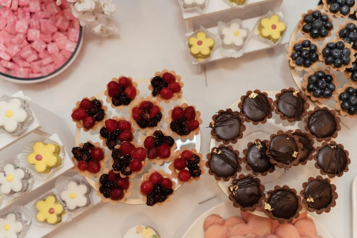 Assortment of decorated desserts including chocolate truffles, berry-topped tartlets, flower-shaped cookies, and pink marshmallows on a white surface. Wedding & Event Planner Hudson Valley, NYC, New York, Bergen County, New Jersey