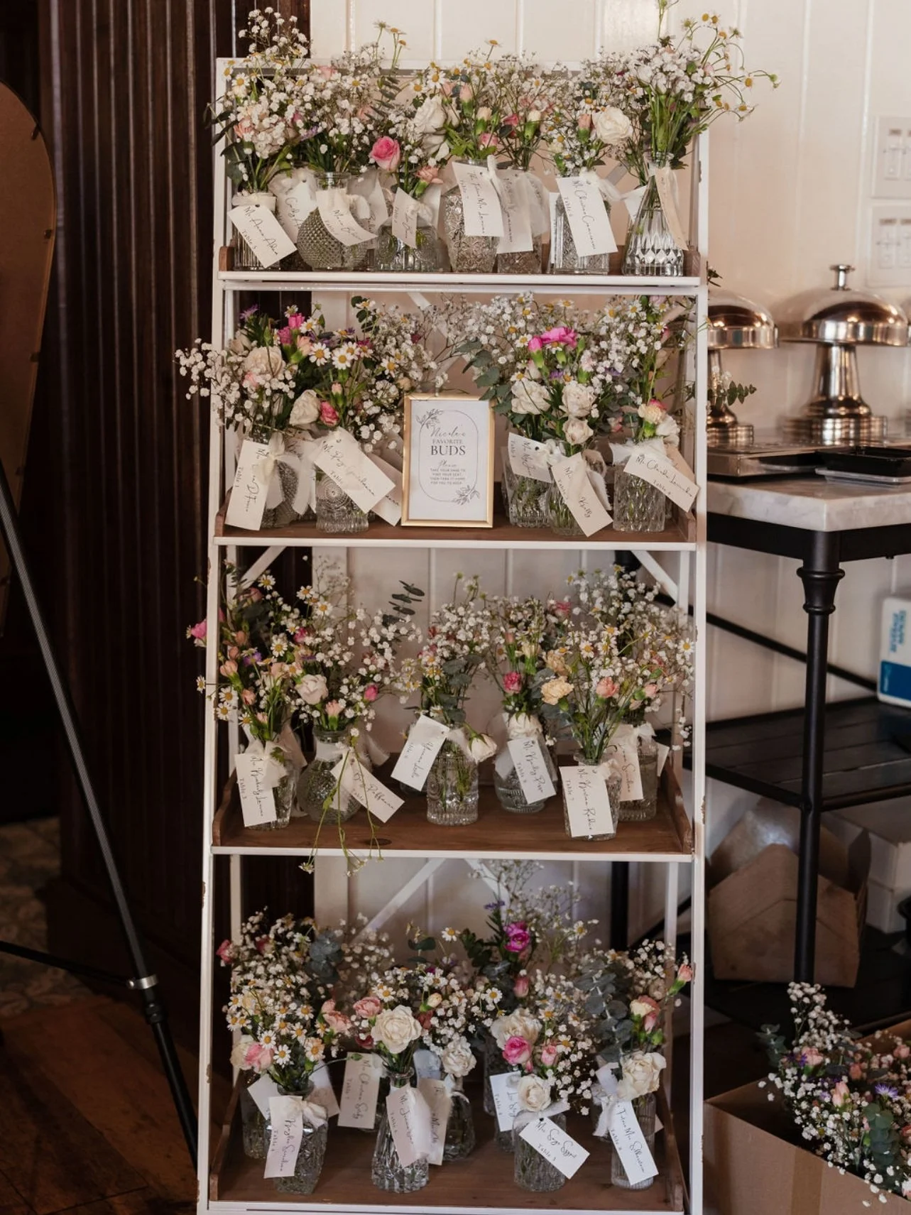 A display shelf filled with various small floral arrangements in glass vases, each with a white tag, and a framed sign reading "Buds" in the center. Wedding & Event Planner Hudson Valley, NYC, New York, Bergen County, New Jersey