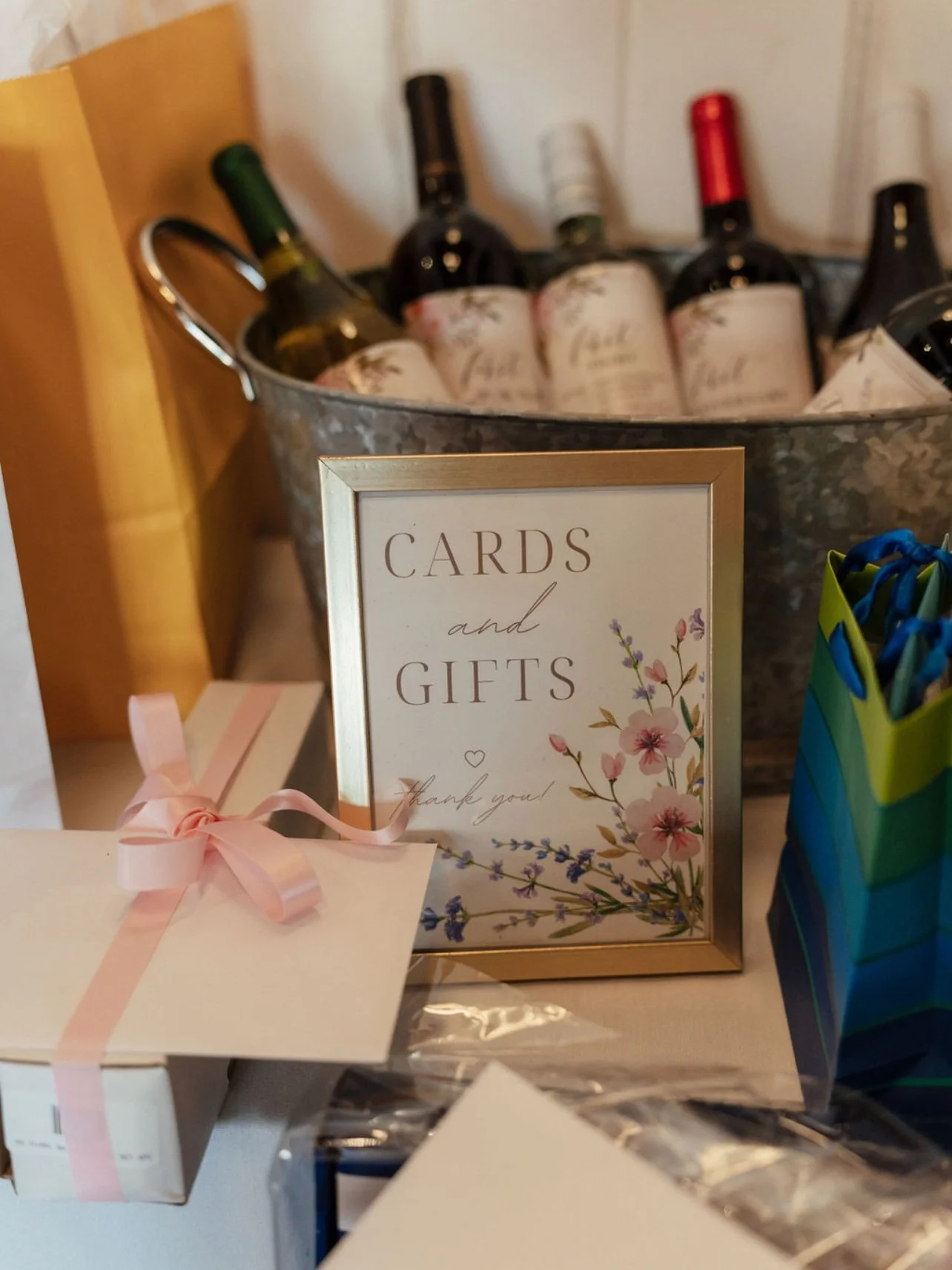 A framed sign that says 'Cards and Gifts, thank you!' with a floral design, a wrapped gift with a pink ribbon, and a colorful striped bag with pens inside. In the background, there is a metal tub filled with bottles of wine. Wedding & Event Planner