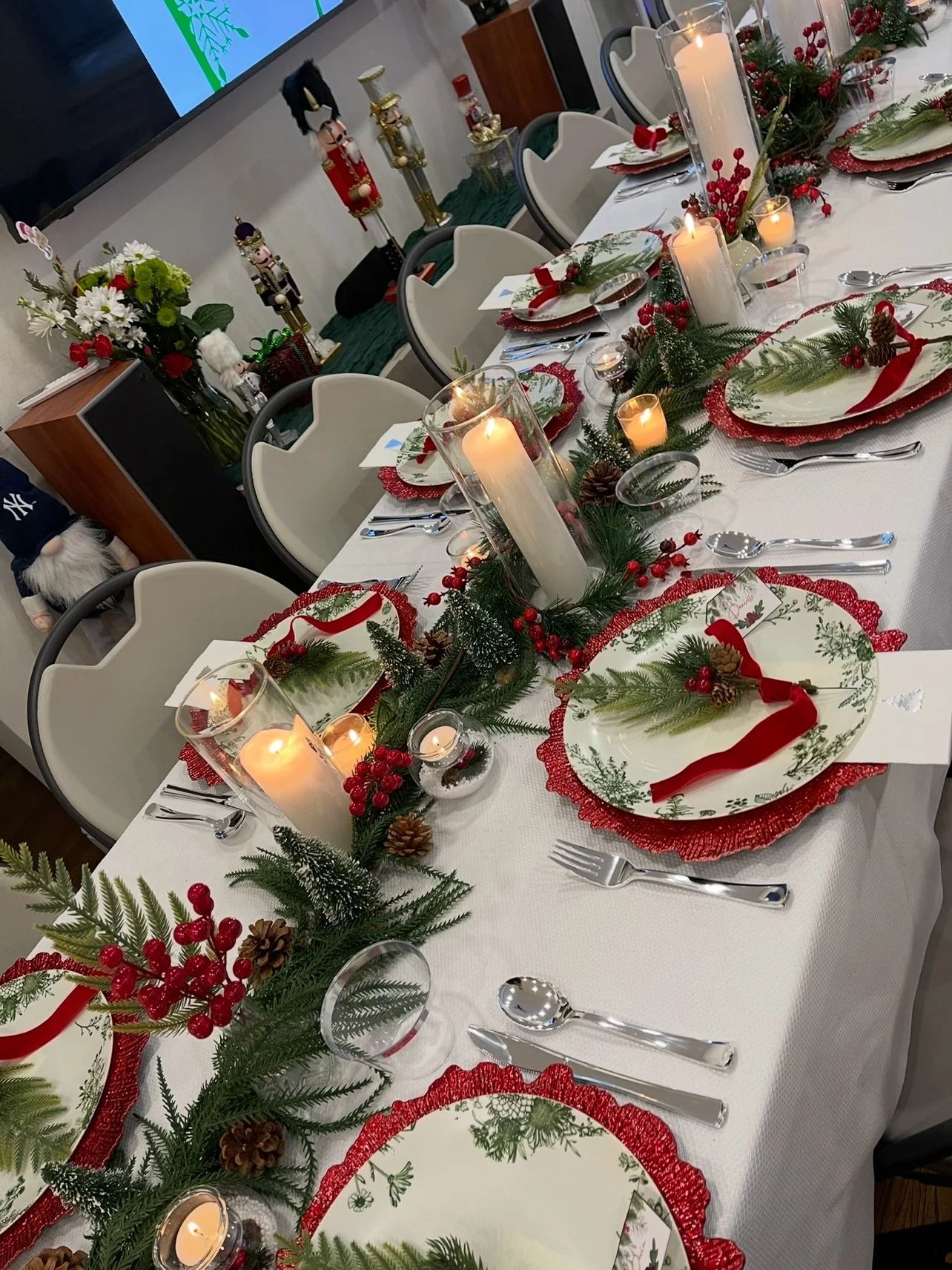 A Christmas dinner table decorated with candles, pine branches, red berries, pinecones, and red ribbons, with place settings on white plates with red and green details. Wedding & Event Planner Hudson Valley, NYC, New York, Bergen County, New Jersey