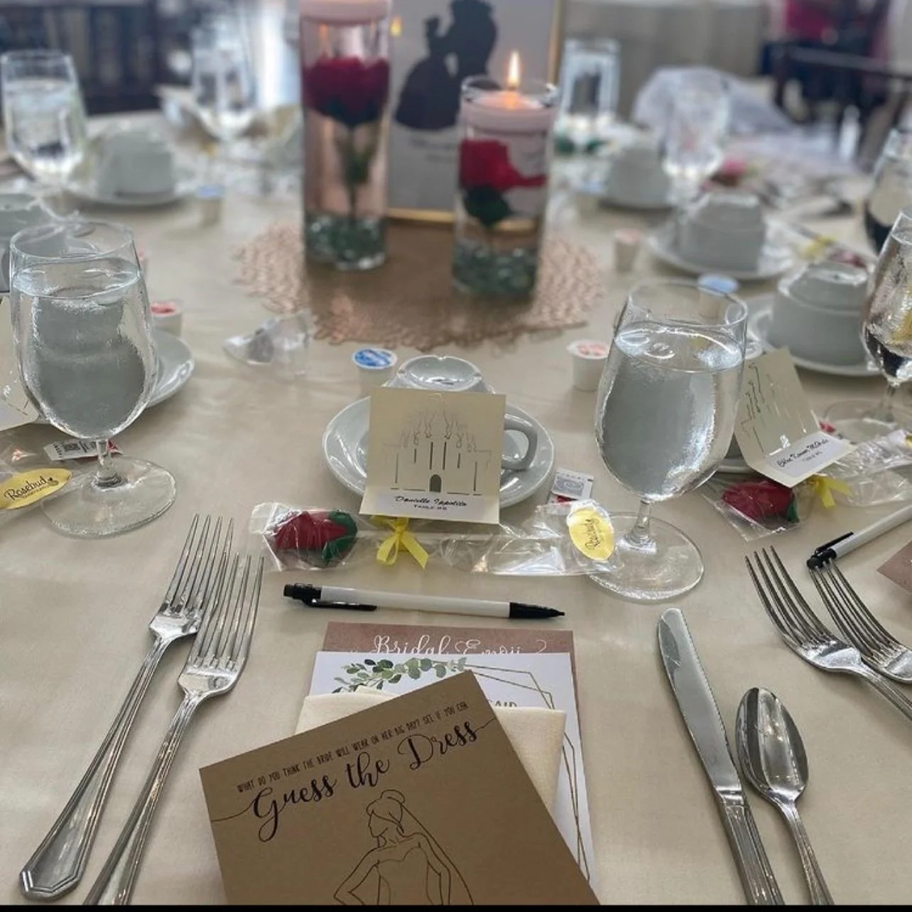Table decorated for a wedding or event with water glasses, silverware, small gift bags, and a 'Guess the Dress' game card, with two tall vases holding roses and candles as centerpieces. Wedding & Event Planner Hudson Valley, NYC, New York, NJ