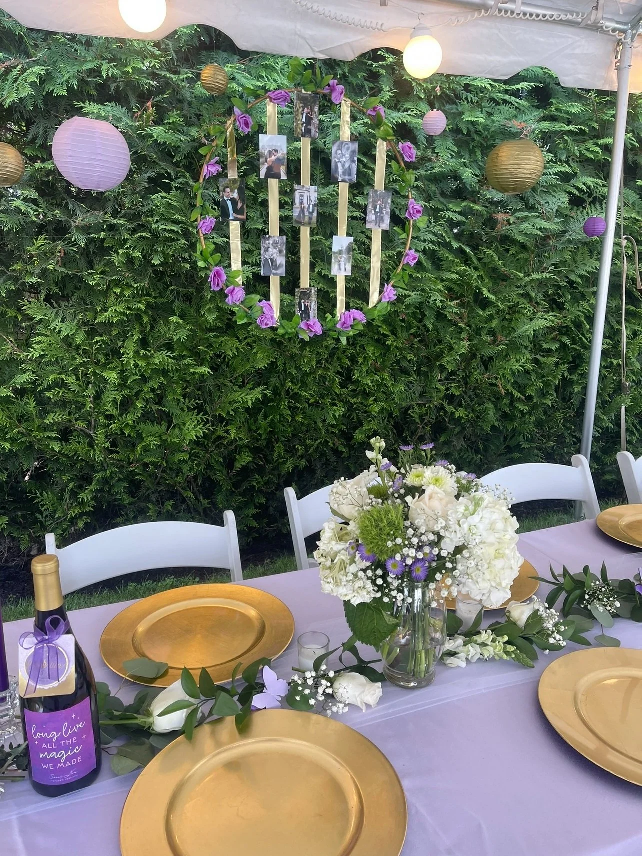 A decorated outdoor table setting with white chairs, gold plates, a large floral centerpiece with white and purple flowers, a bottle of pink bubbly with a purple ribbon, and a photo display decorated with purple flowers and photos hanging on a circul