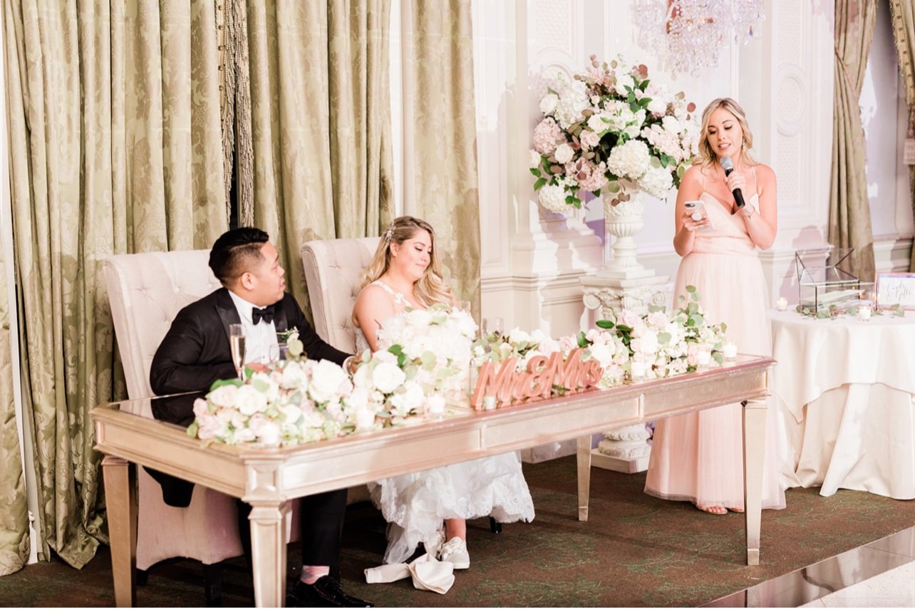 A wedding reception with a bride and groom sitting at a decorated table, and a woman giving a speech with a microphone, floral arrangements, and elegant curtains in the background.