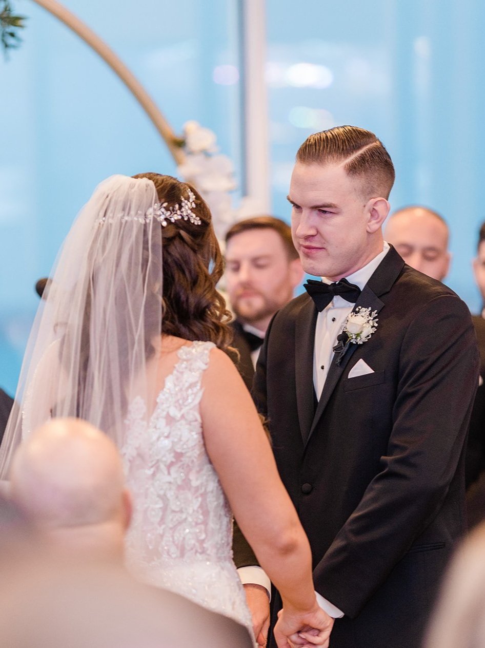 A bride and groom holding hands during their wedding ceremony, facing each other, with a blue background and ceremony guests behind them.