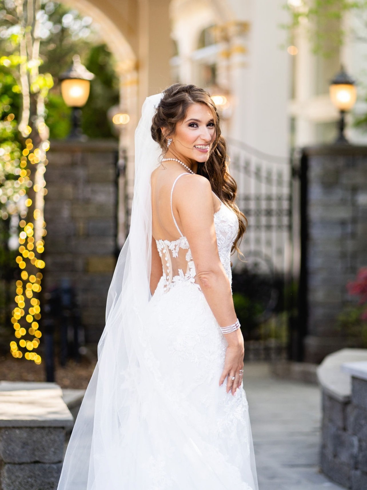 A bride in a white wedding dress with lace detailing, posing outdoors with a smile, holding her hand near her hip, and wearing a veil and pearl jewelry. In the background, there are stone walls, decorative lights, and greenery.