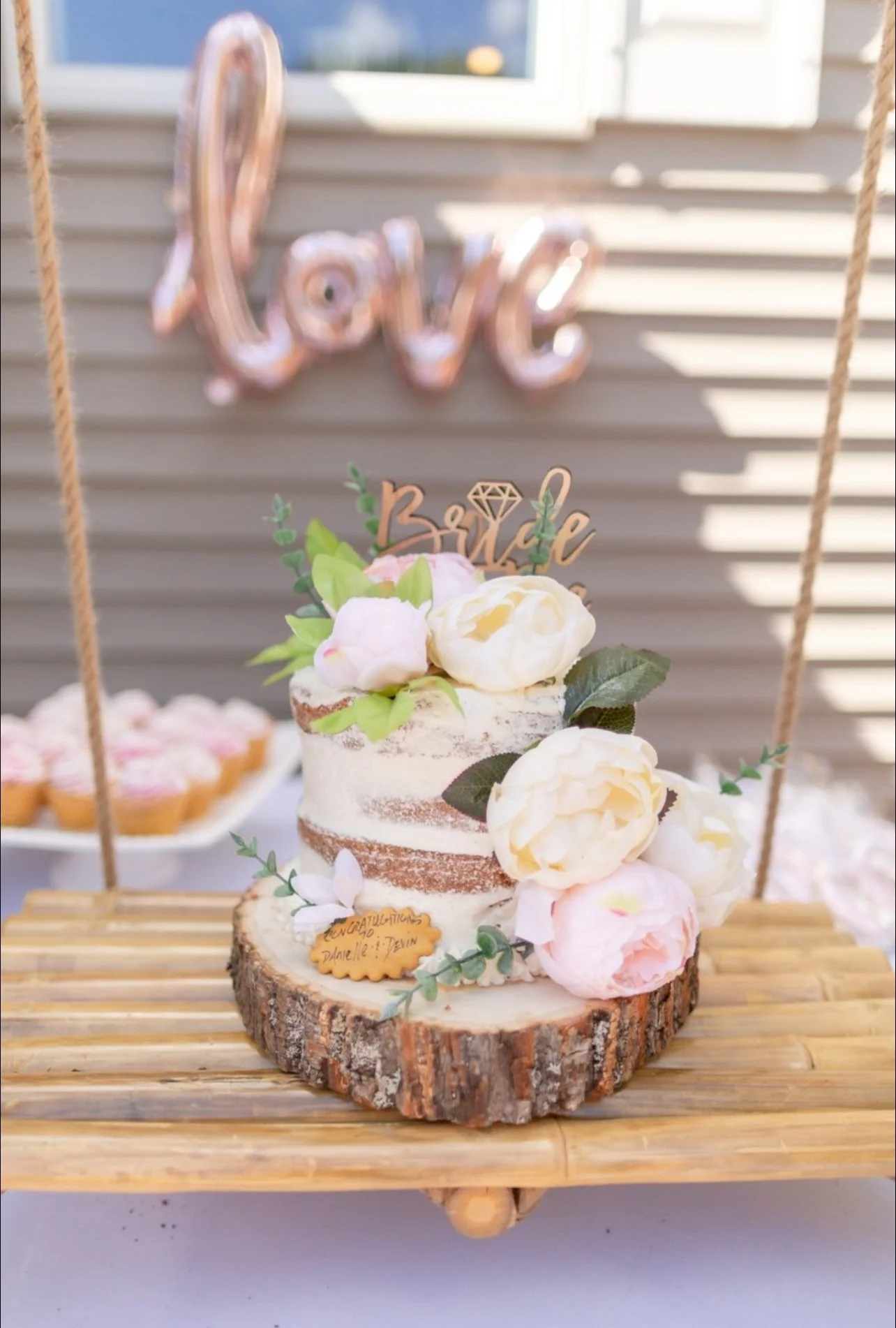 A small rustic wedding cake decorated with white and pink flowers and greenery, sitting on a wooden slab on a bamboo table. In the background, there is a blurred metallic letter sign that reads 'love' and a plate of cupcakes.