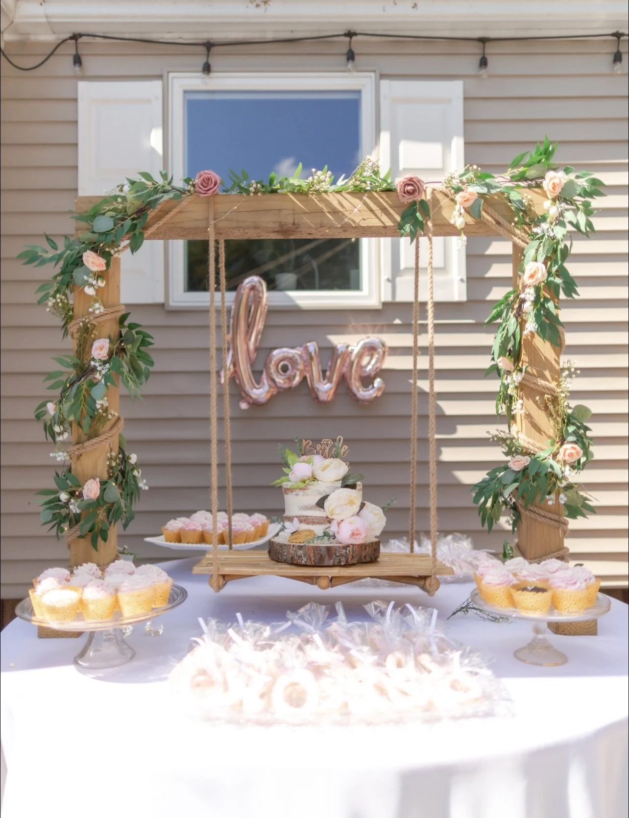 A dessert table with a rustic wooden swing decorated with pink and white roses and greenery, featuring a pink "love" balloon and a small cake with flowers on a wooden slab, surrounded by cupcakes and cookies, set against a beige house exterior.