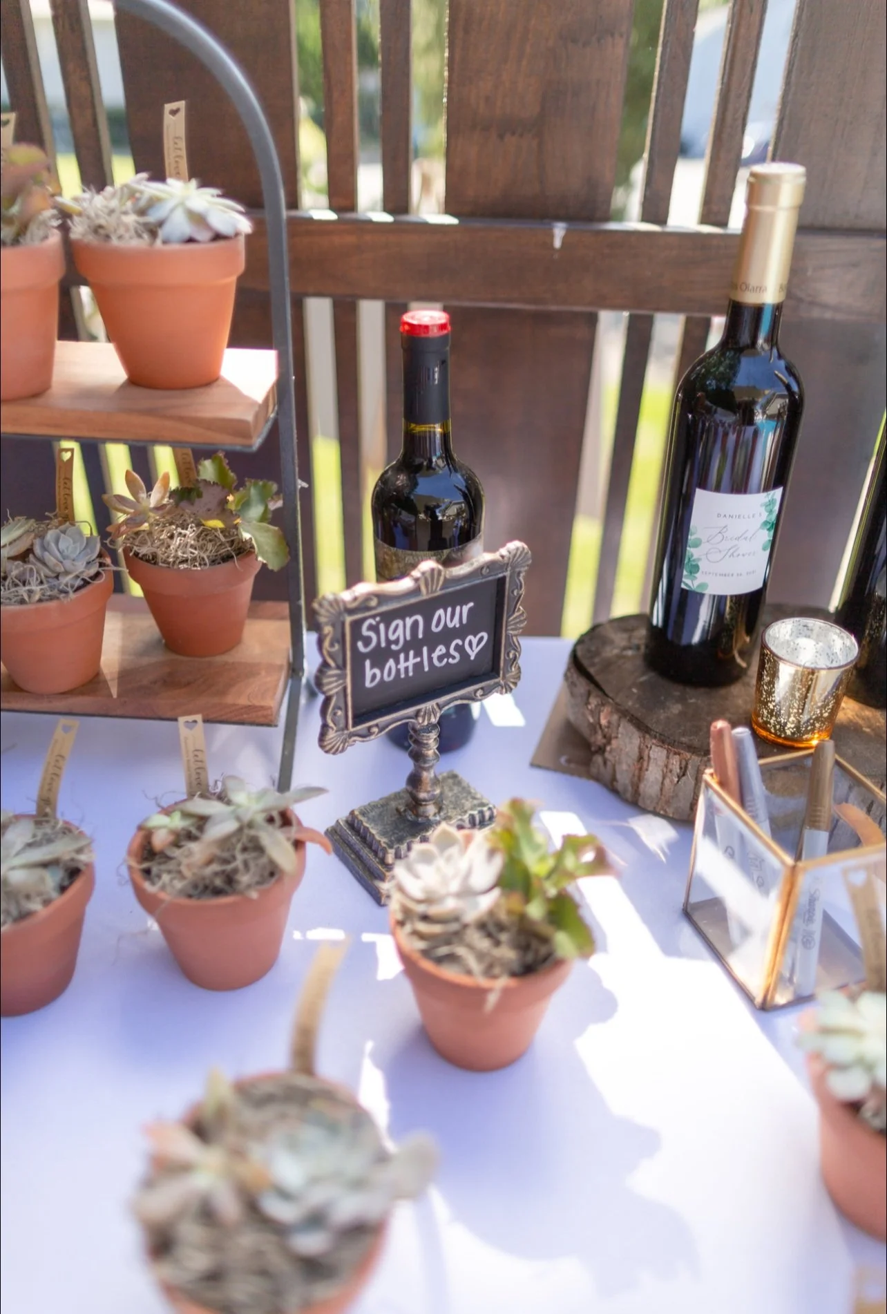 Table with potted succulents, bottles of wine, a sign that reads 'Sign our bottle,' and decorative items at an outdoor event.