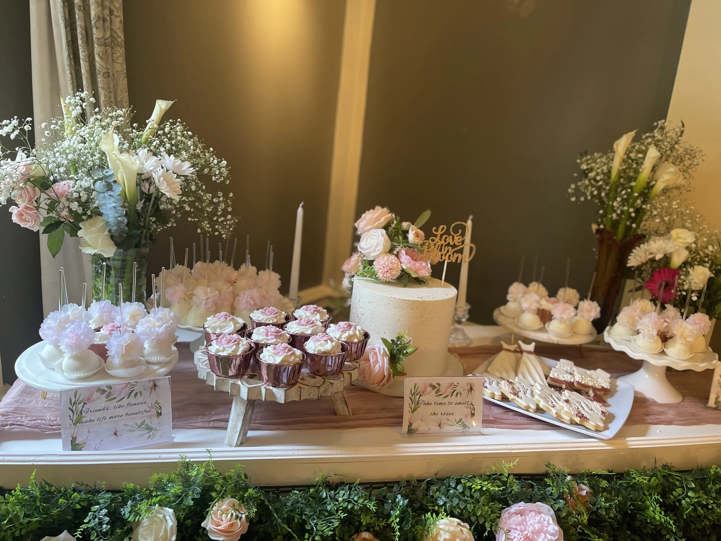 Elegant dessert table with pink and white cupcakes, floral arrangements, and a white cake with pink flowers, decorated with signs and surrounded by greenery. Wedding & Event Planner Hudson Valley, NYC, New York, Bergen County, New Jersey