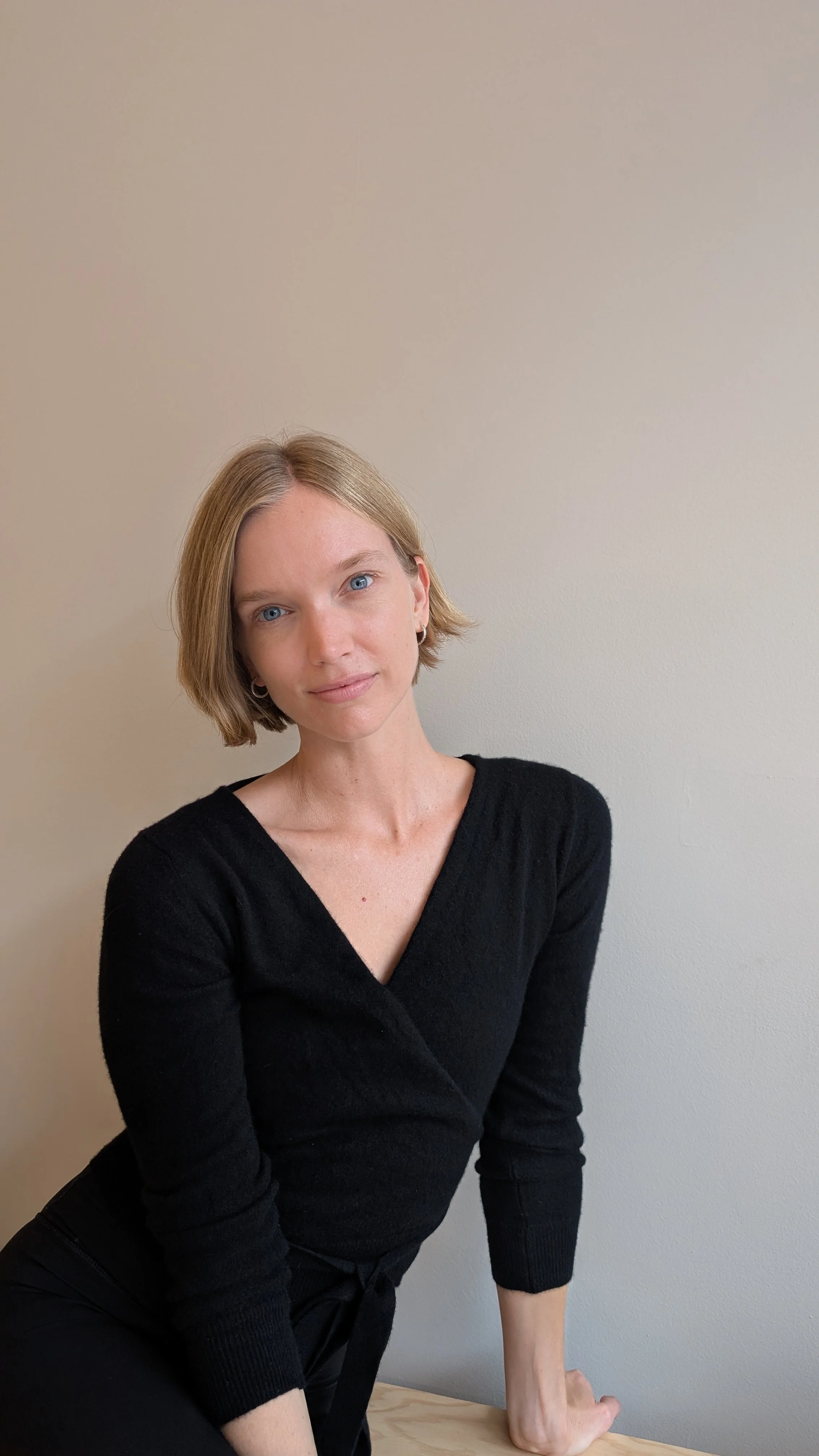 A woman with short, blonde hair and blue eyes taking a selfie in a room with beige walls and a framed picture in the background.