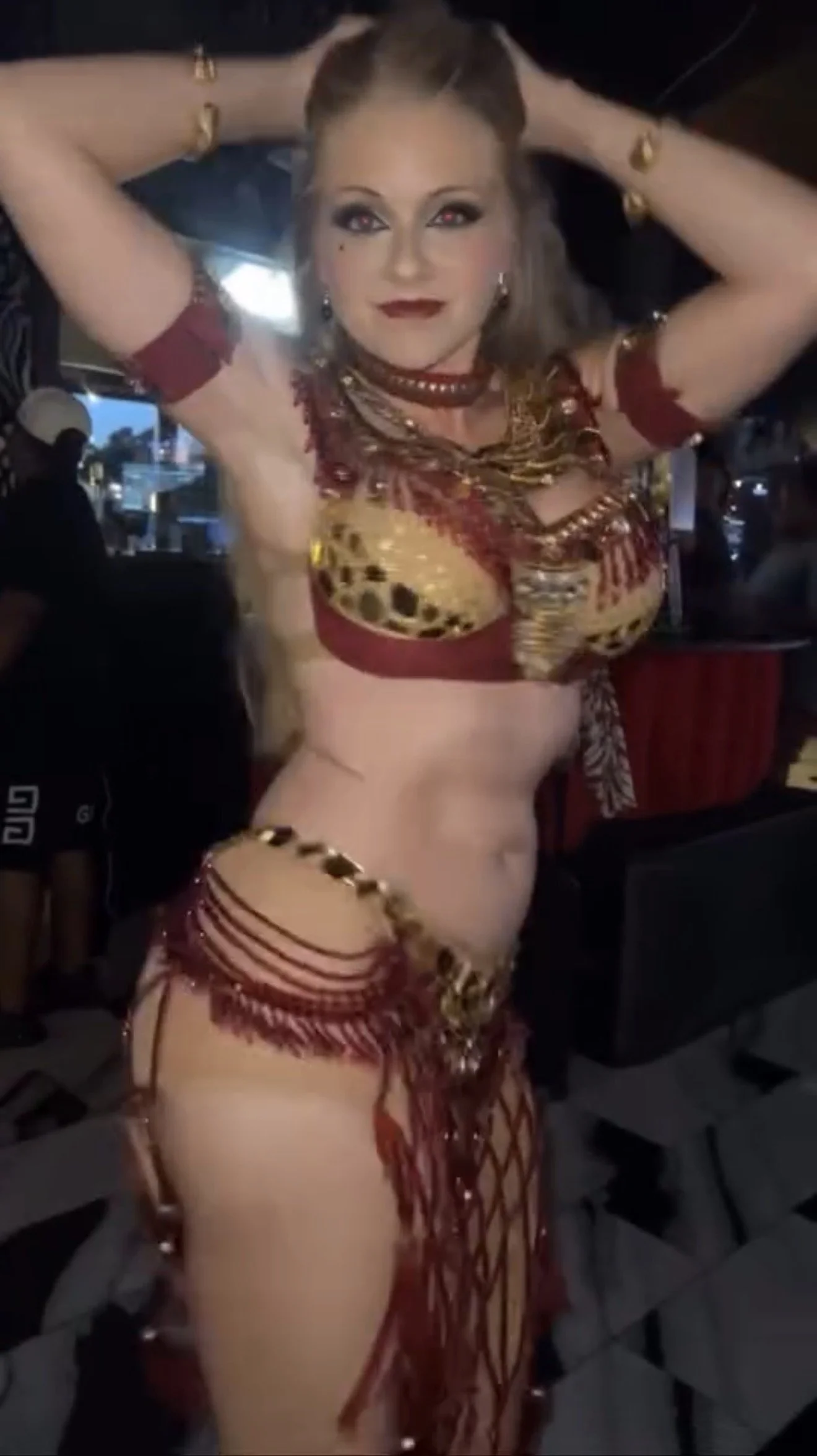 Woman in a red and gold belly dance costume with jewelry, posing indoors.