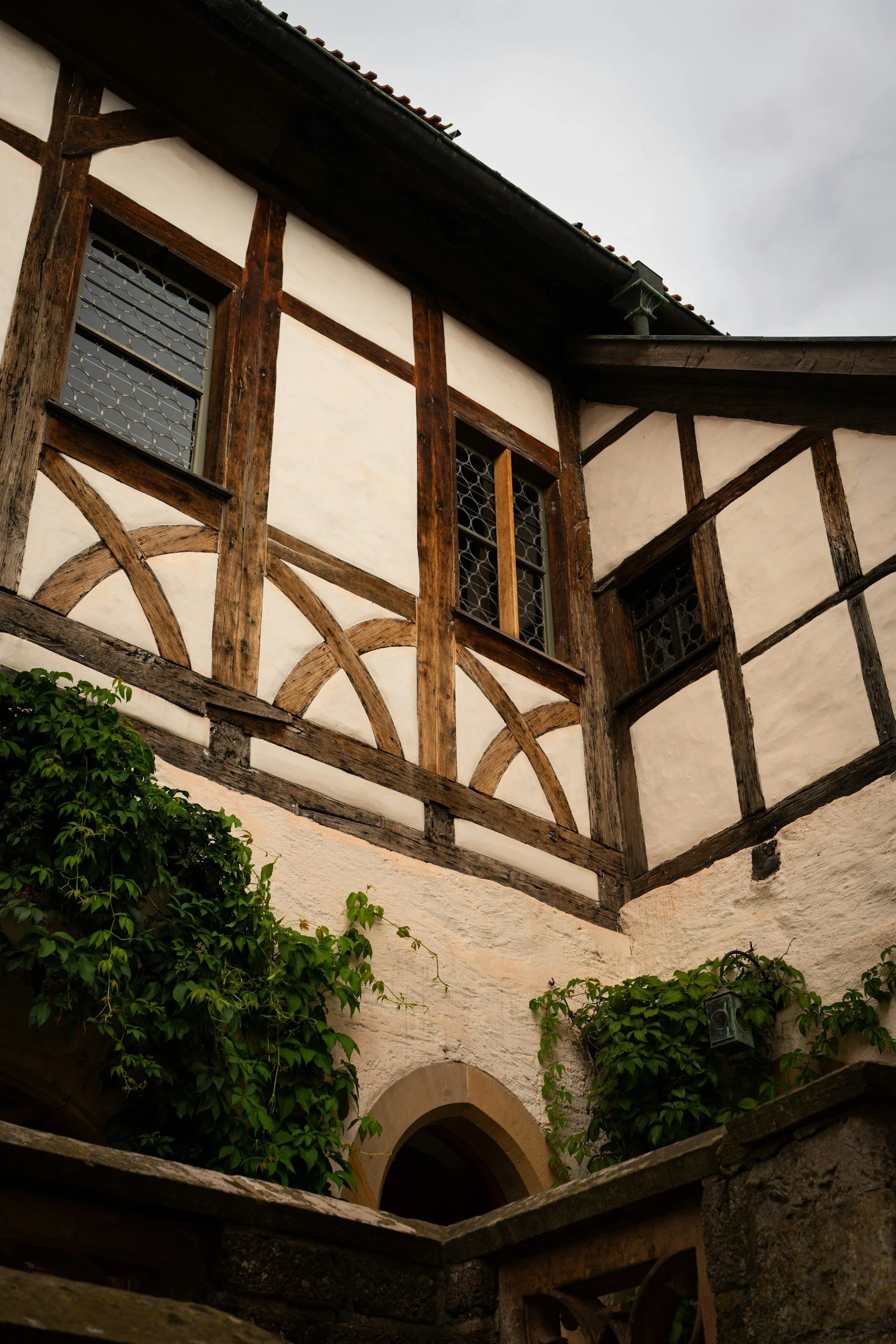 A traditional European-style building with whitewashed stucco walls, dark wooden beams, and lattice windows, surrounded by green foliage and ivy.