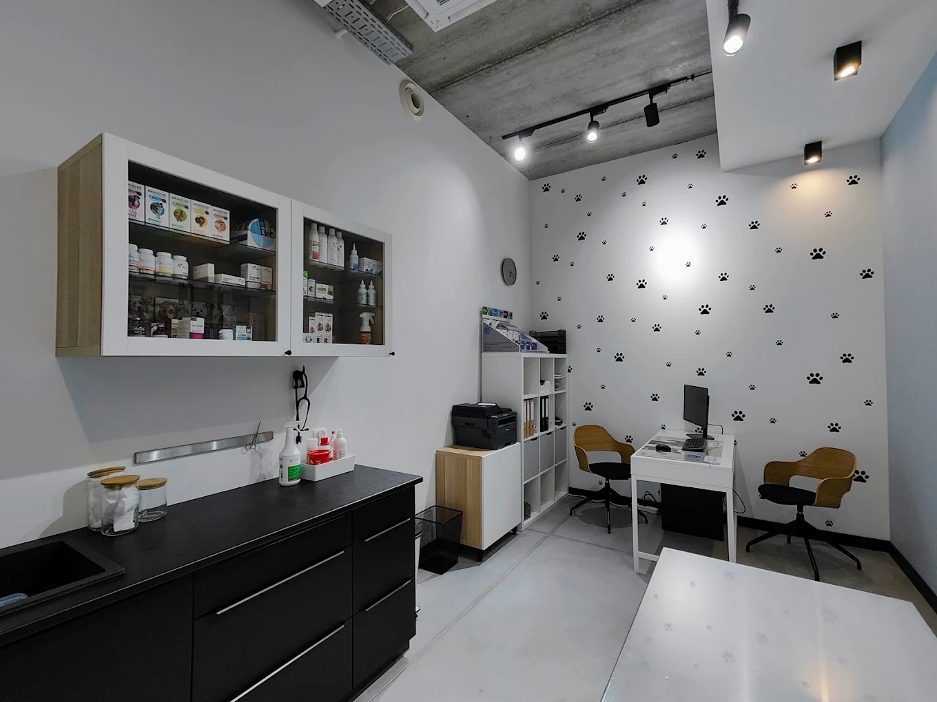 Gabinet weterynaryjny z białymi ścianami ozdobionymi czarnymi naklejkami z odciskami łap. Stoją tam dwa drewniane krzesła, małe białe biurko z komputerem i biała półka z artykułami biurowymi. Widoczna jest również czarna szafka z lekami.