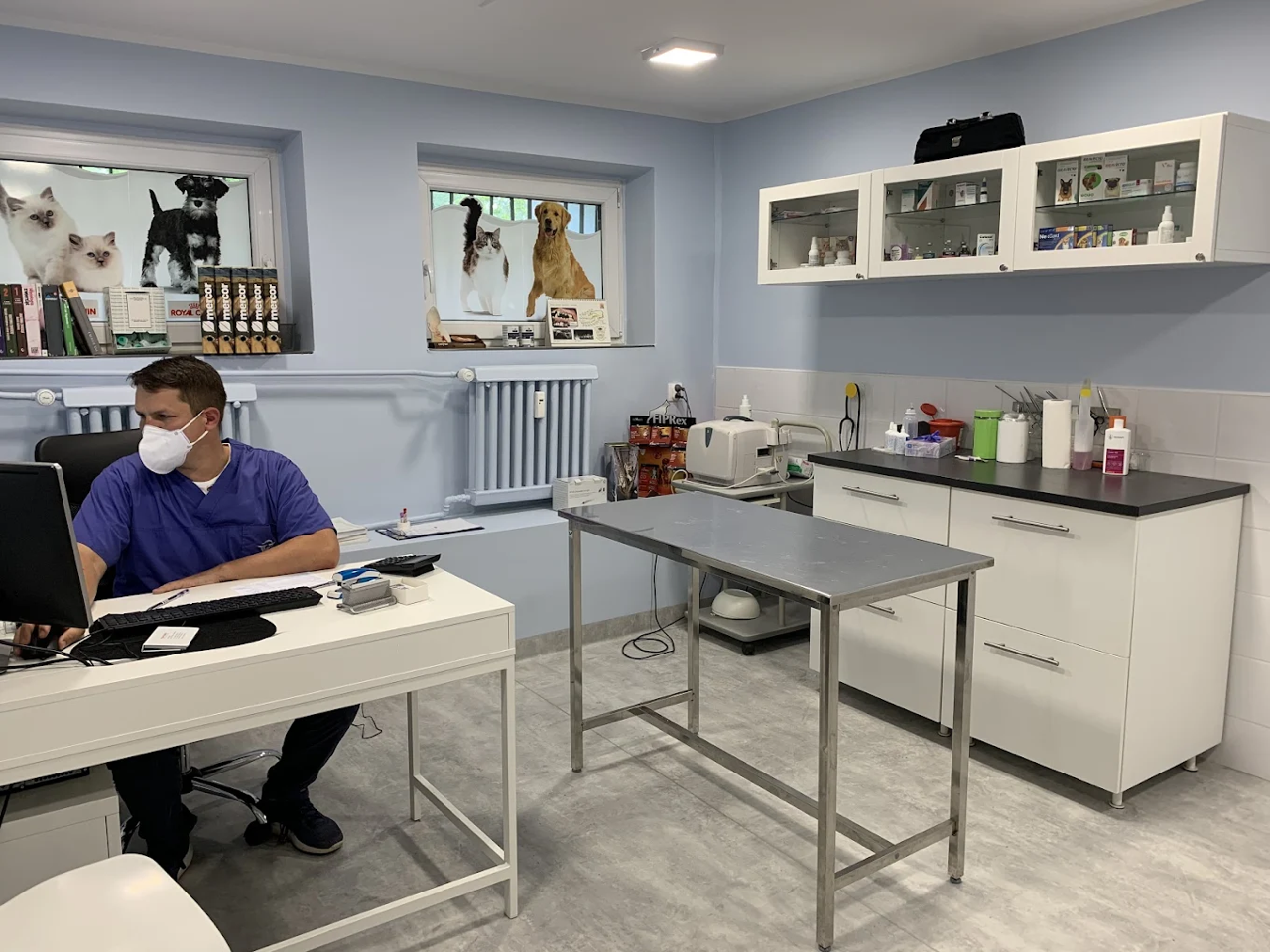 Gabinet weterynaryjny z pracownikiem ubranym w fartuch i maskę, komputerem, środkami farmaceutycznymi, stołem do badań ze stali nierdzewnej i zdjęciami zwierząt w oknach.