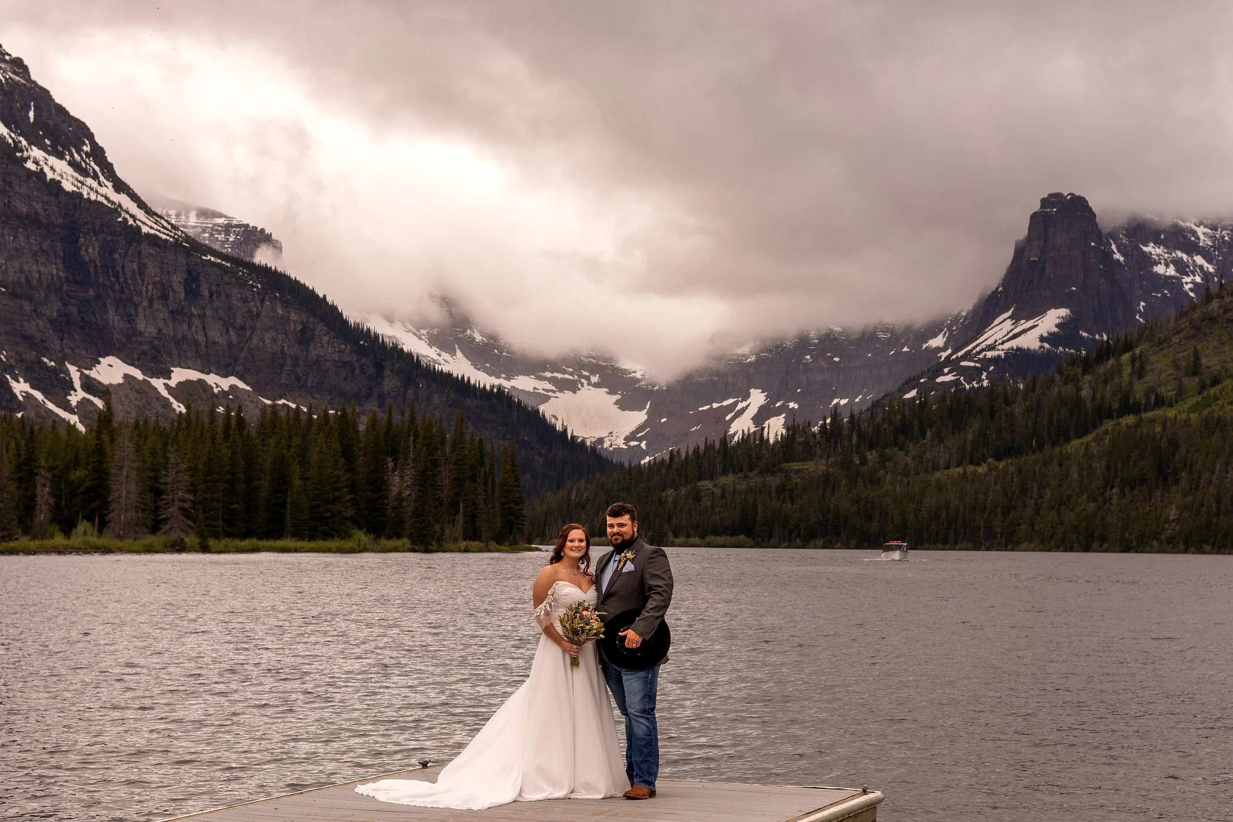 A couple in wedding attire standing on a dock by a lake, with mountains and forest in the background, cloudy sky overhead in Glacier National Park.