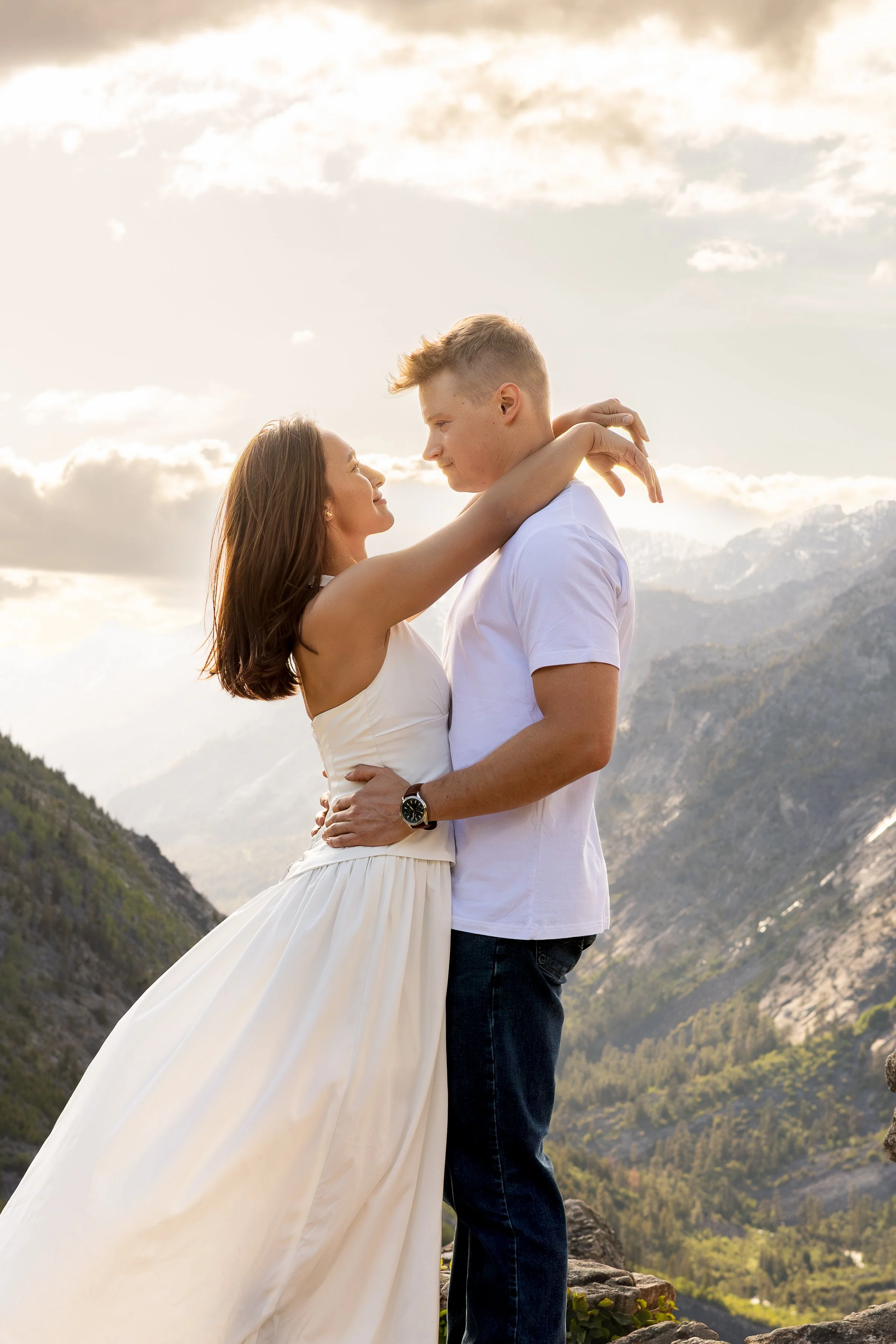 A couple standing very close — possibly embracing — outdoors in a mountainous area with clouds in the sky, gazing into each other's eyes.