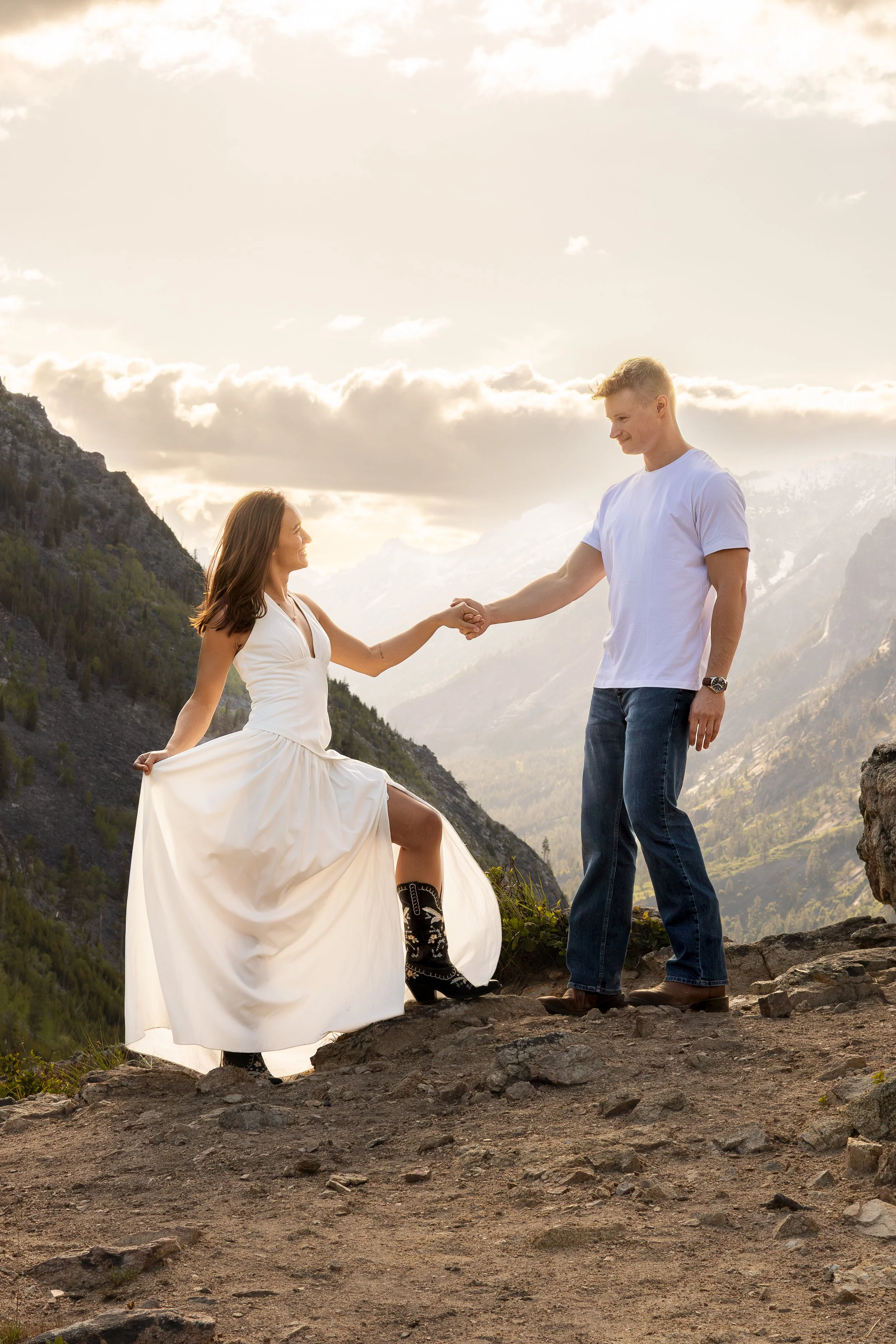 A woman in a white dress and cowboy boots sitting on a rock, holding hands with a man in a white T-shirt and jeans, standing on a rugged mountain landscape during sunset.