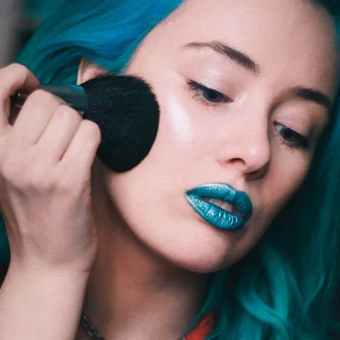 Femme aux cheveux bleus appliquant du maquillage sur son visage.