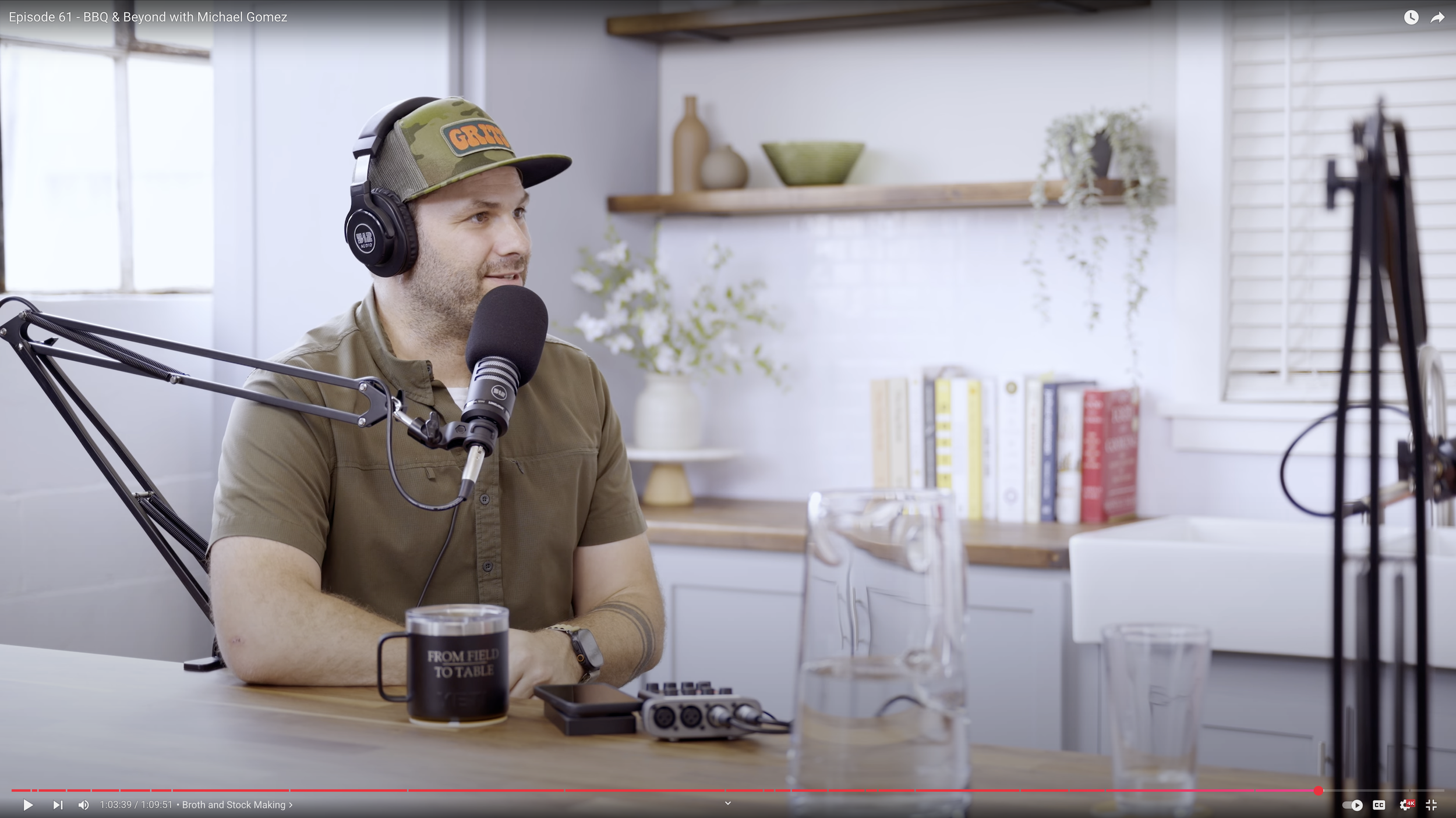 A man wearing headphones and a baseball cap with a microphone, sitting at a table with a coffee mug, glass of water, and podcast recording equipment, in a cozy, well-lit room with bookshelves and decorative plants.