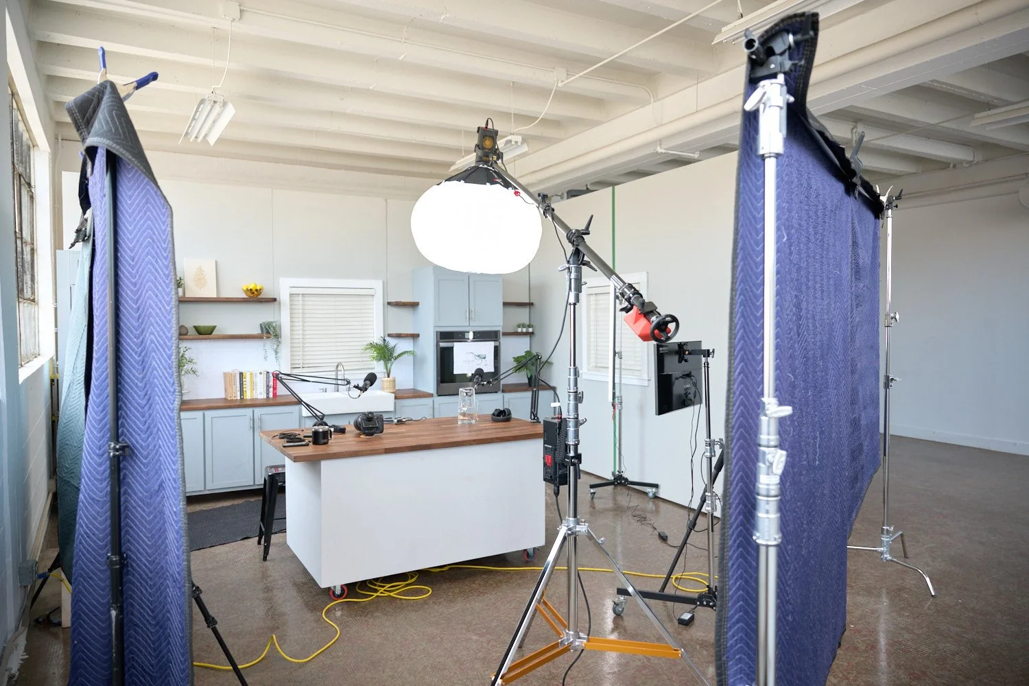 A professional photography studio set with lighting equipment, backdrop, and a white kitchen counter with shelves in the background.