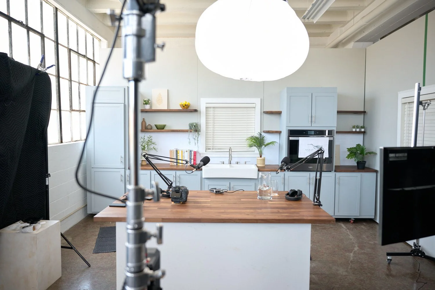 A kitchen set up for a podcast or video recording with cameras, microphones, and lights. The kitchen features light blue cabinets, a white farmhouse sink, and wooden shelves with decorative items and plants. There is a large round light fixture hangi
