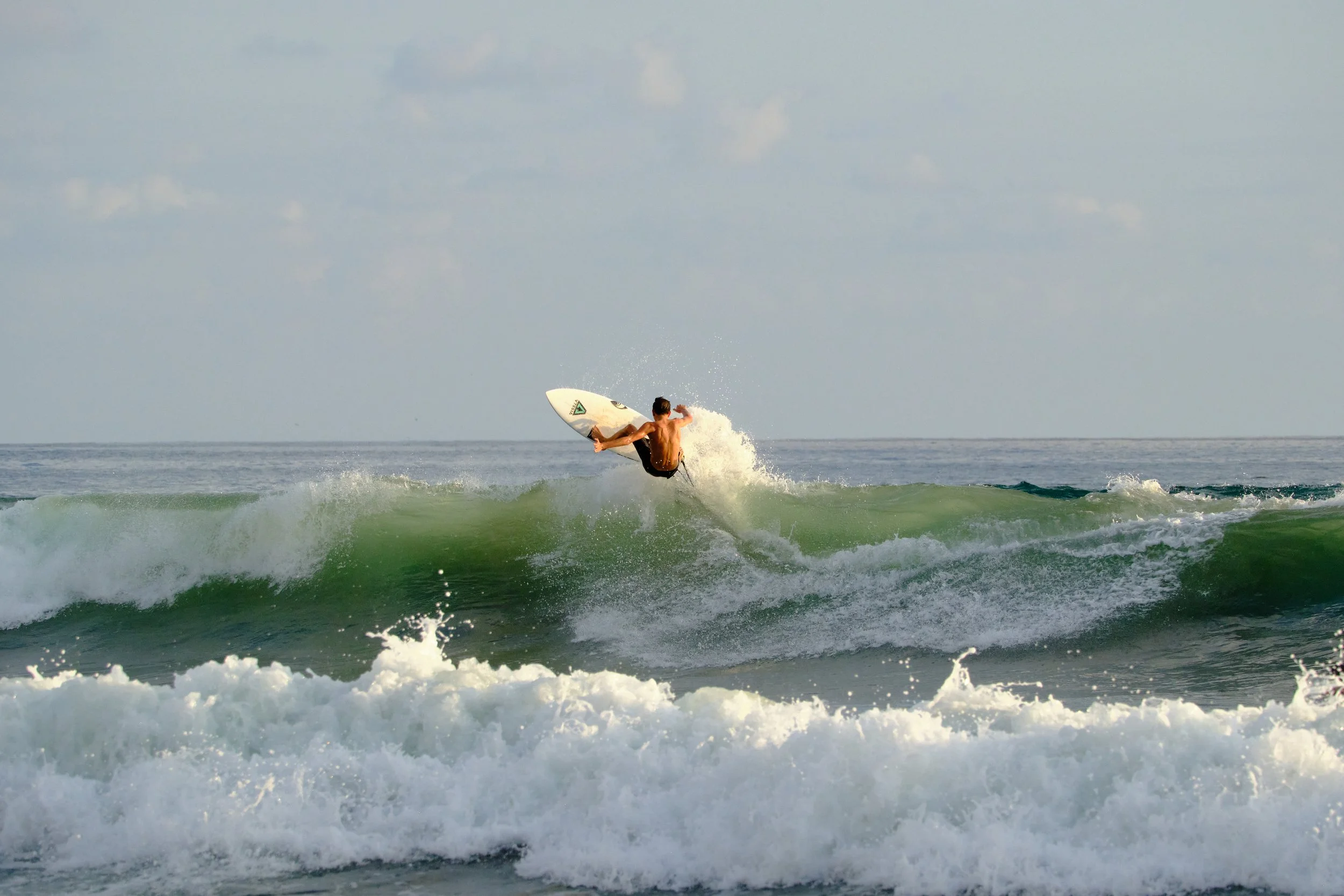 A surfer performs an aerial maneuver on a wave in the ocean under a partly cloudy sky.