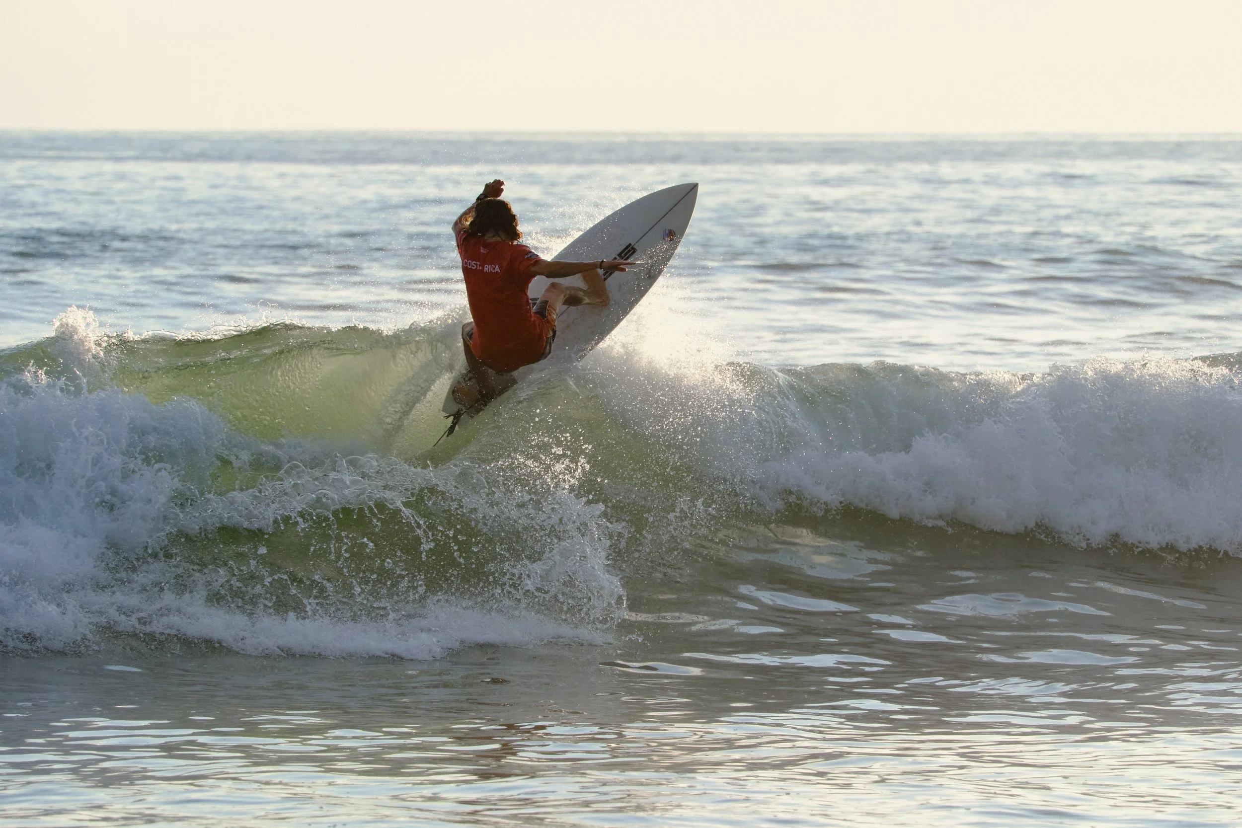 A person surfing on a wave in the ocean, wearing an orange shirt and shorts, with sunlight reflecting on the water.