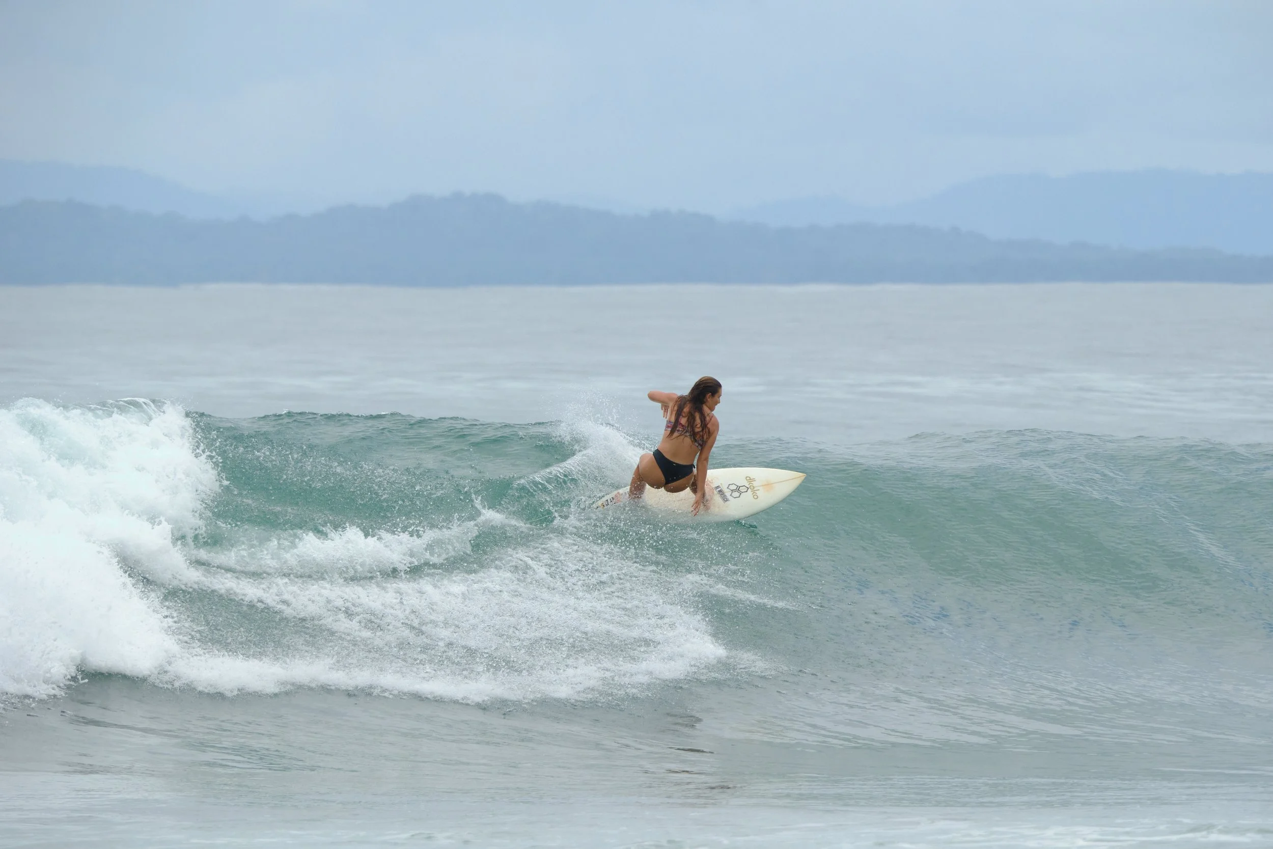 Woman surfing on a wave in the ocean with mountains in the background.