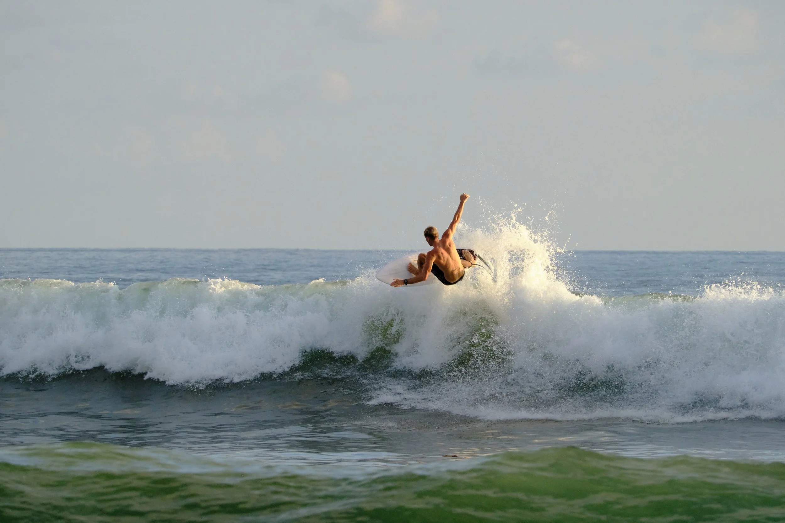 A shirtless man surfing on a wave in the ocean, jumping into the air with a surfboard.