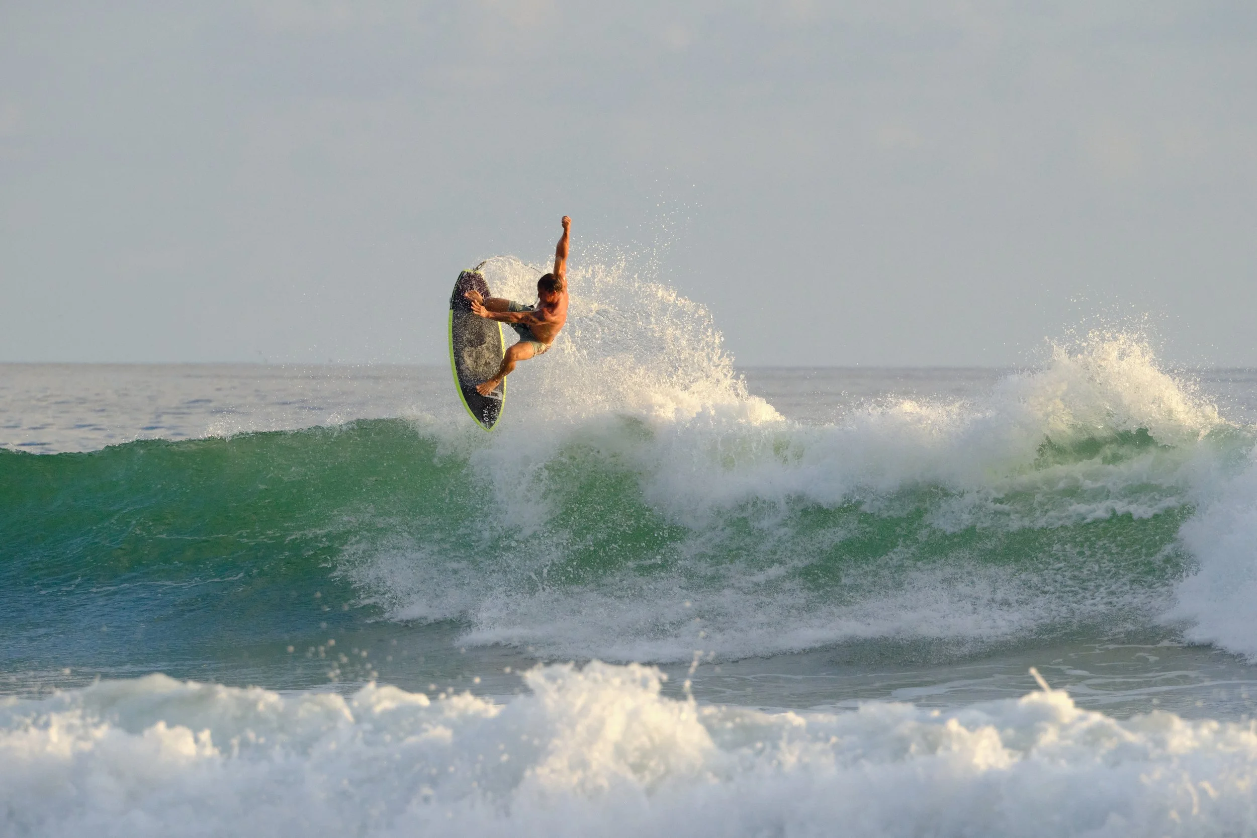 A male surfer in a tan swimsuit performing an aerial trick on a green wave at the beach.