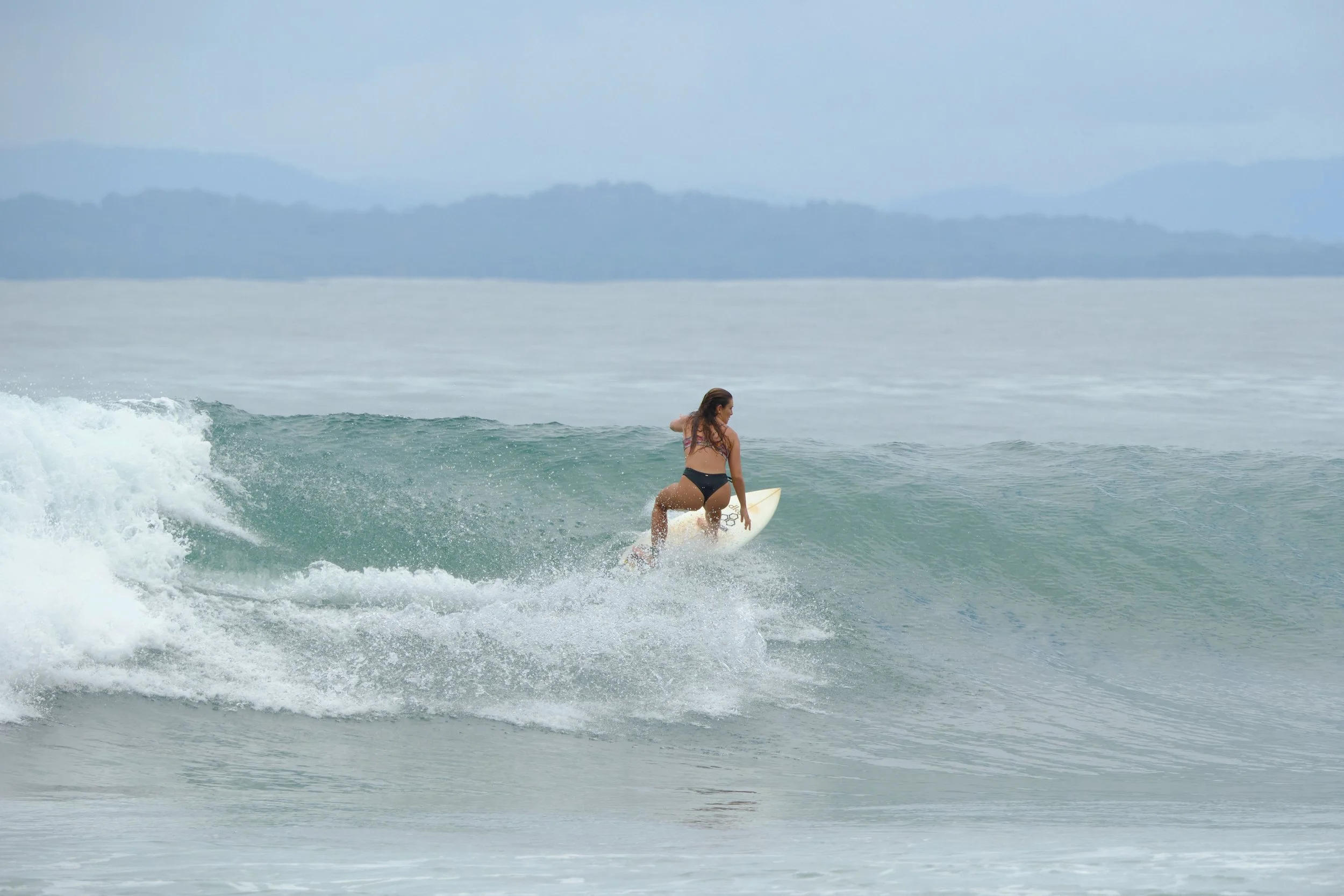 A woman surfing on a wave in the ocean, wearing a black bikini with a scenic mountainous background.