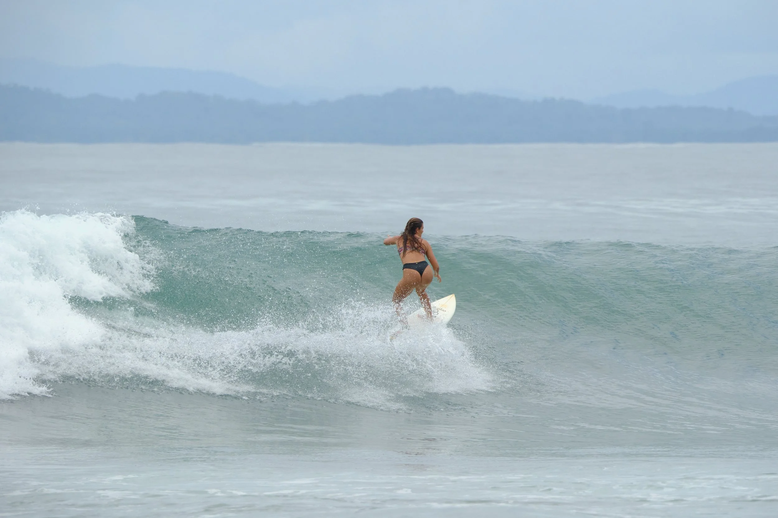 A woman in a bikini surfing on a small wave in the ocean with distant mountains in the background.