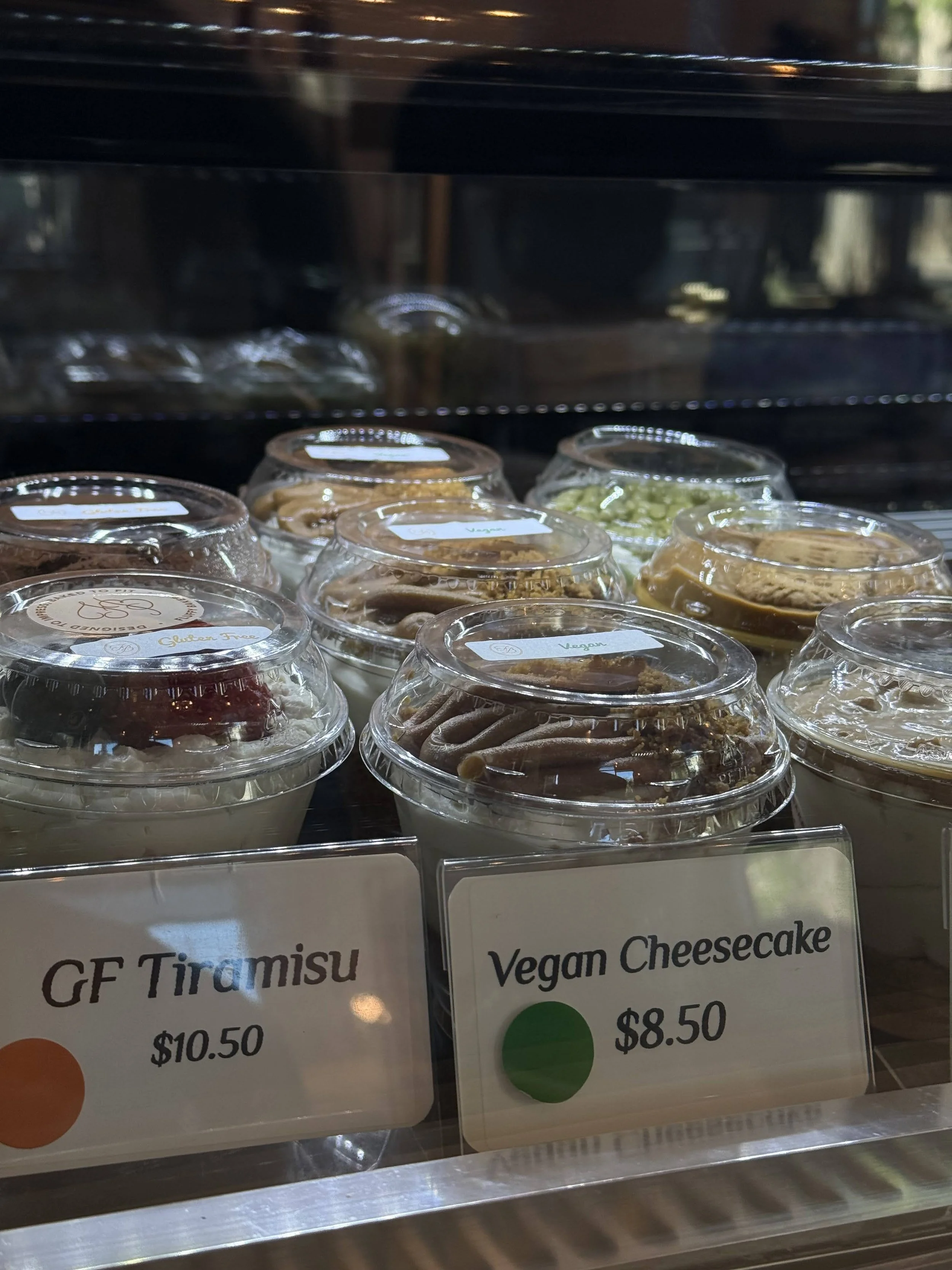 Clear plastic containers of vegan cheesecakes on display, with signs reading 'GF Tiramisu' and 'Vegan Cheesecake' in front.
