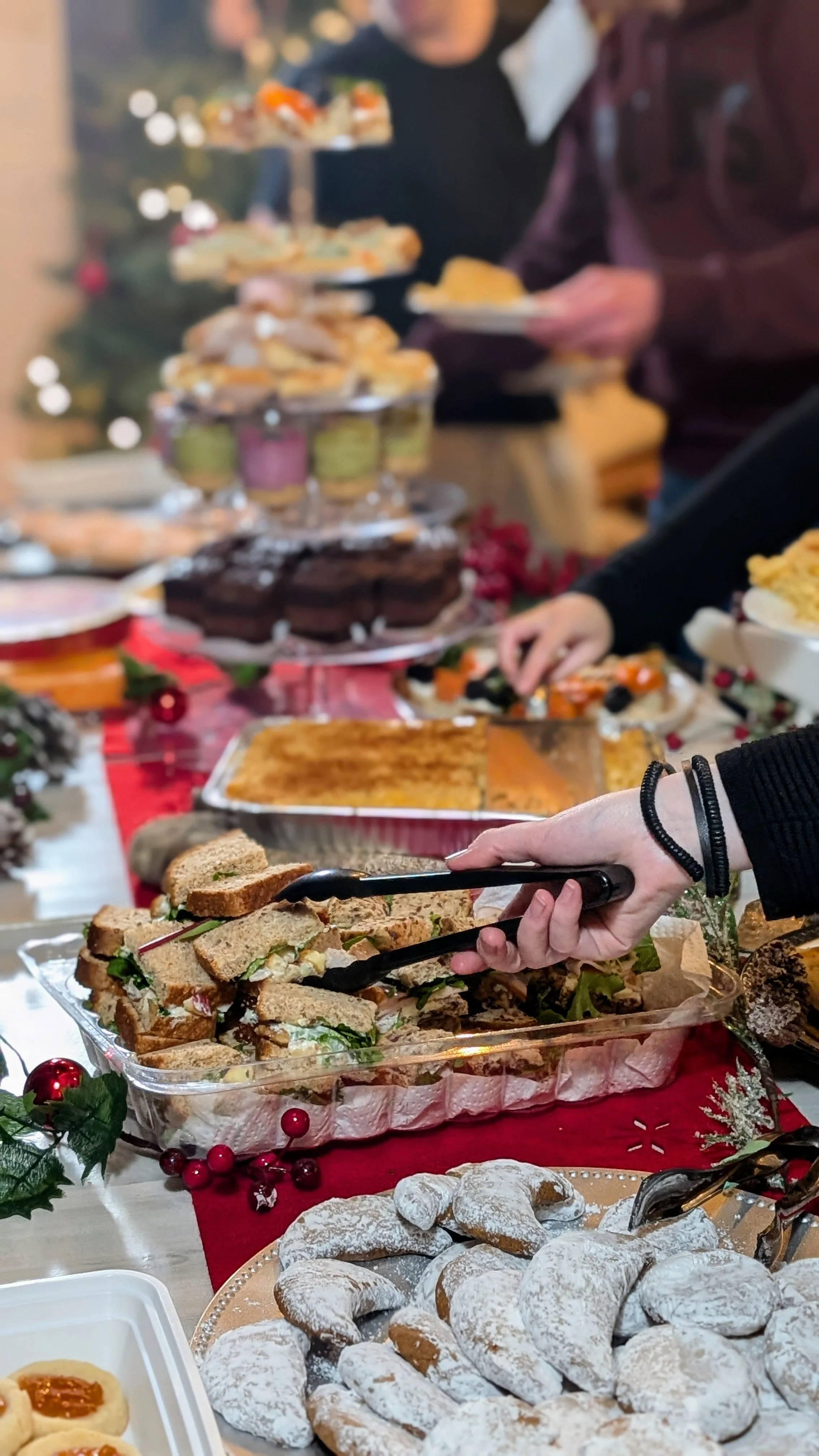 A festive holiday buffet table with various desserts and finger foods, including powdered sugar-covered cookies, sandwiches, cakes, and pastries, decorated with holly and red berries.