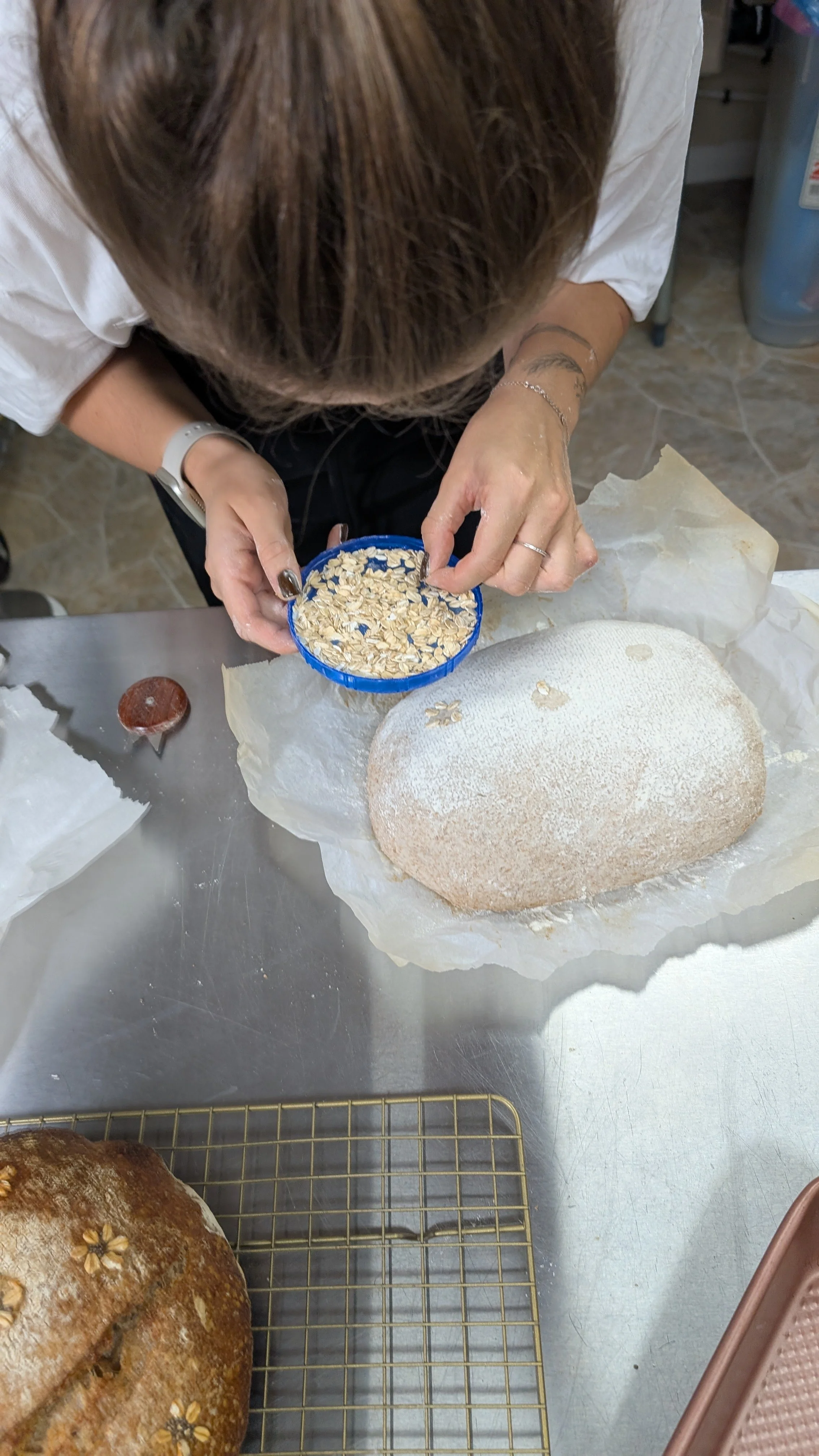 A person decorating a bread loaf with oats, standing over a metallic table with white parchment paper, other baked breads, and baking tools.