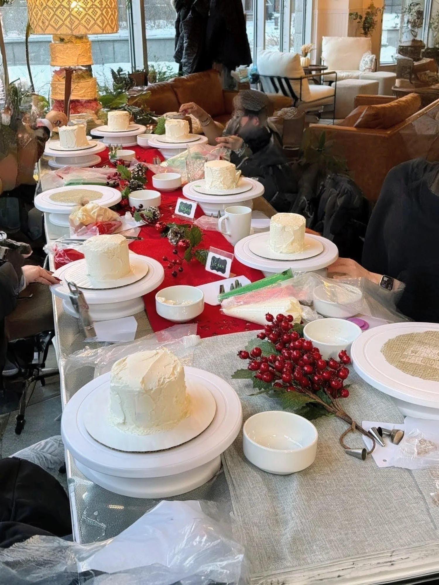 A long dining table decorated with red and gray table runners, topped with small cakes, bowls, berries, and some tables with unused plates and utensils. People are gathered around the table, setting or preparing for a celebration in a cozy, well-lit room with large windows and comfortable seating.