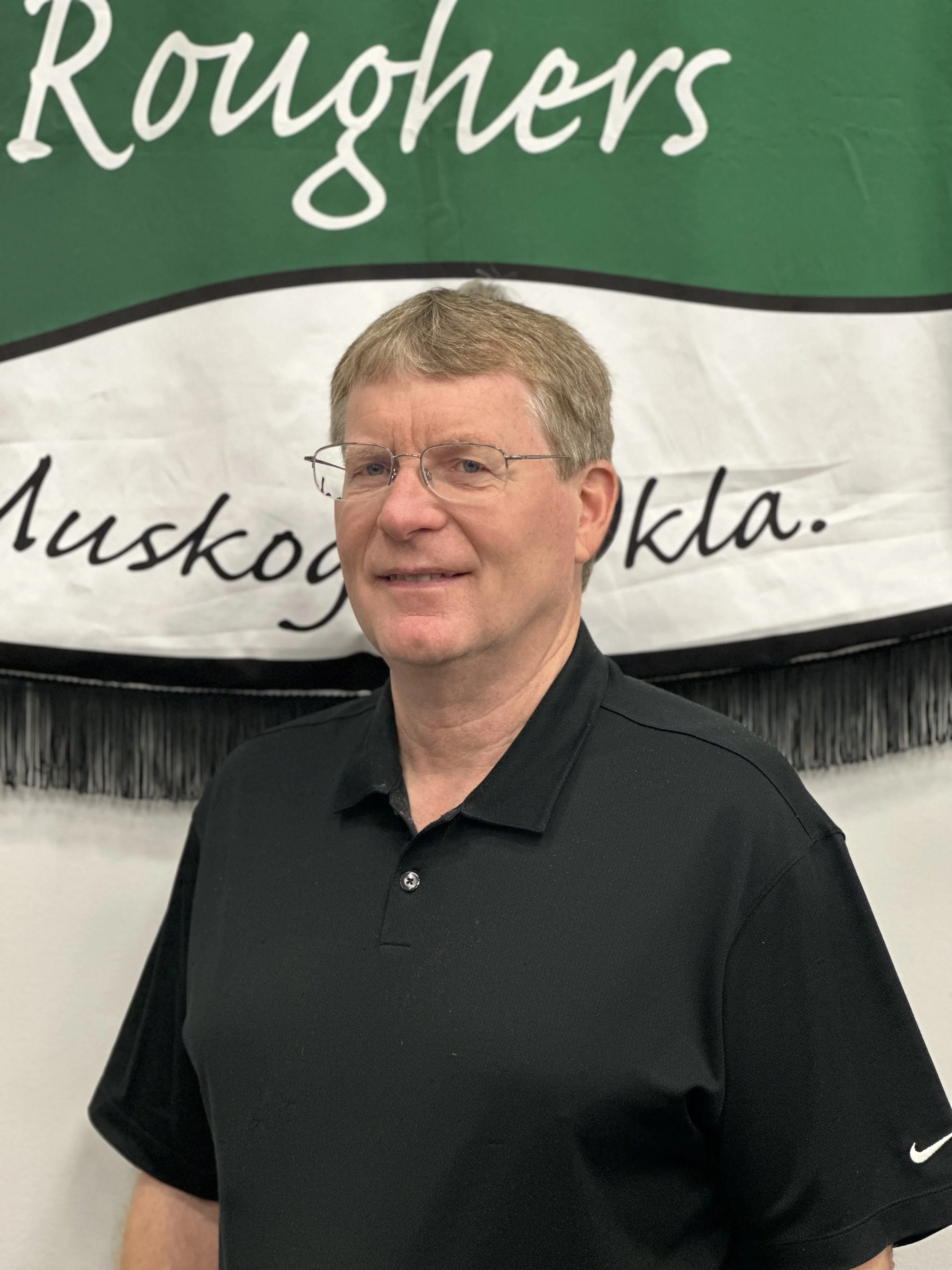 A man with glasses and short light brown hair, wearing a black polo shirt, standing in front of a banner that reads 'Roughers'.