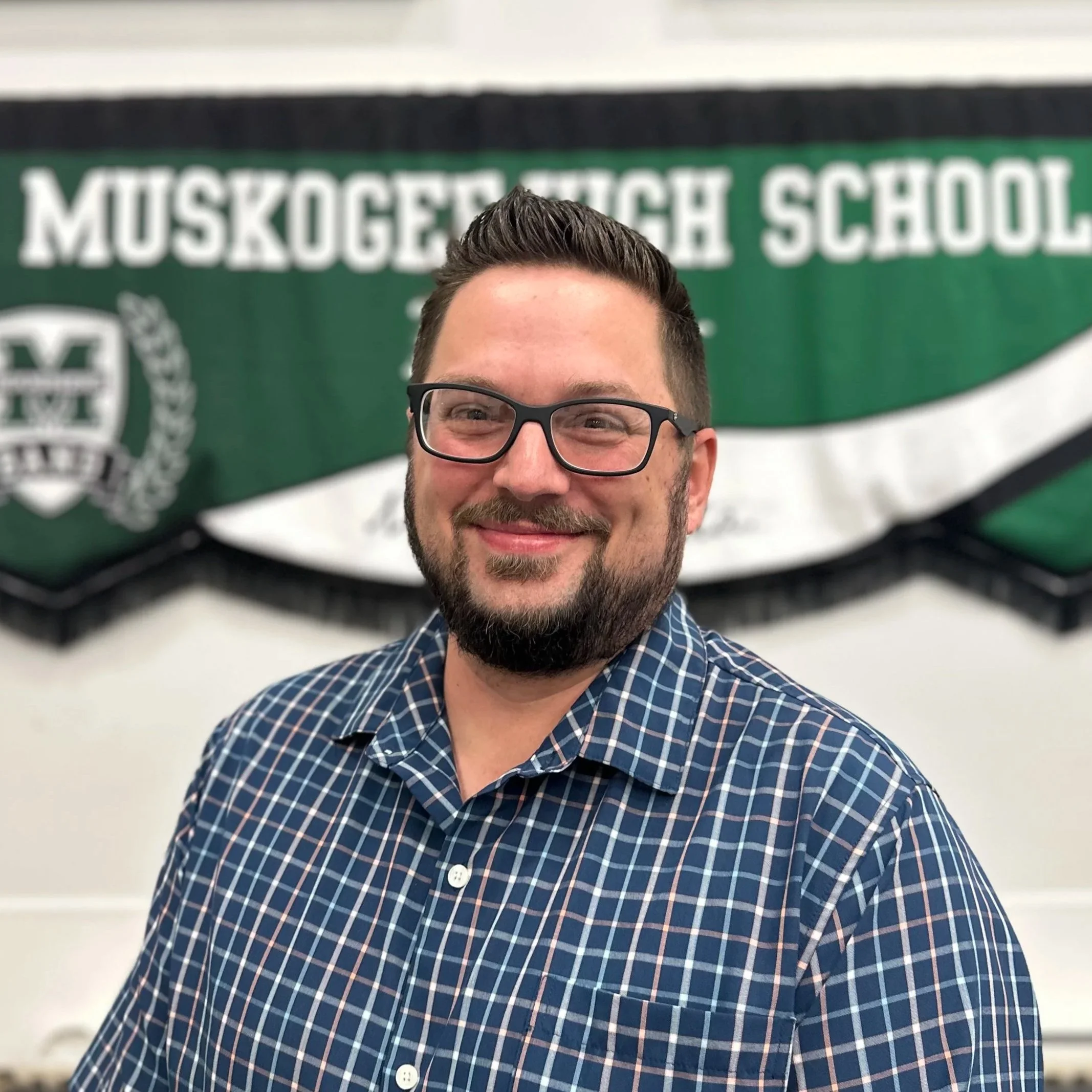 A man with glasses and a beard smiling in front of a green and white Muskegon High School banner.