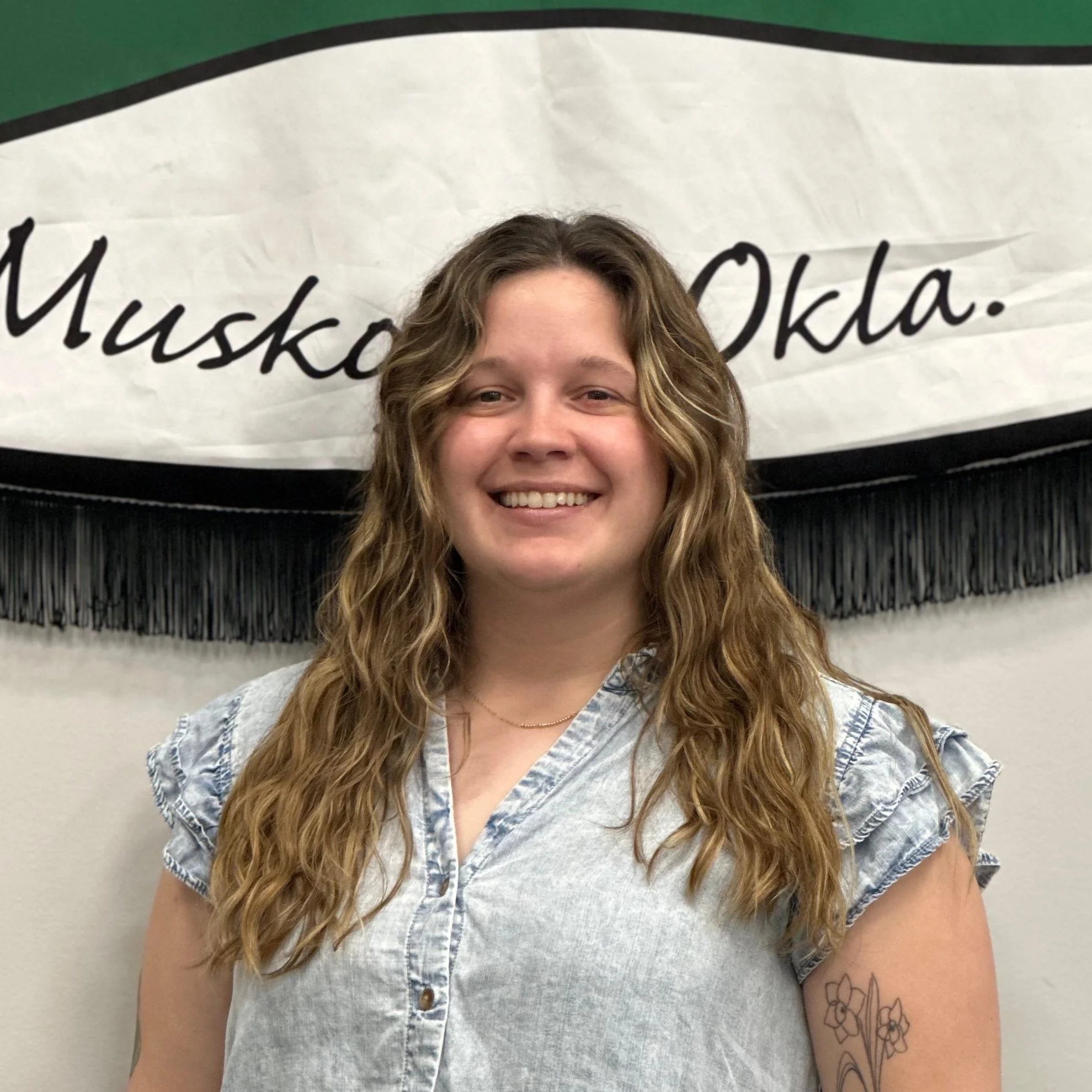 A young woman with long, wavy hair smiling at the camera, standing in front of a white and black Oklahoma-themed banner.