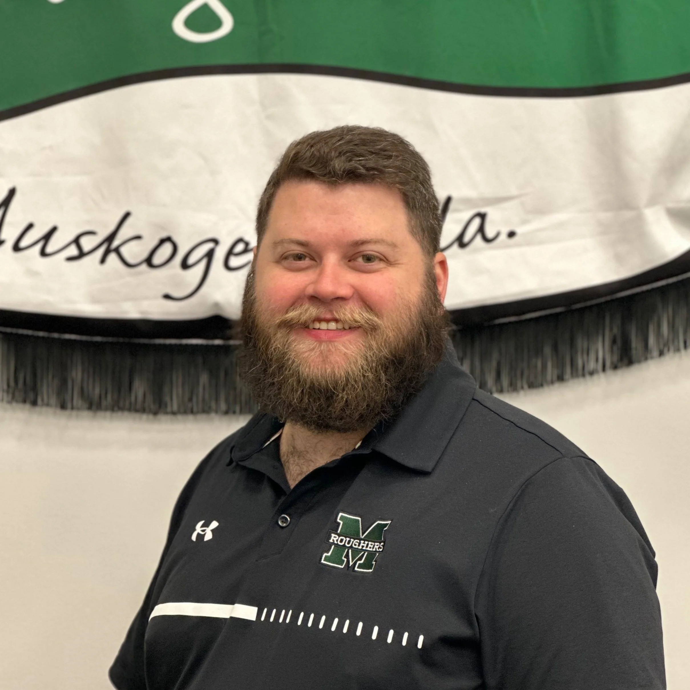 A man with a beard and short hair smiling, wearing a black polo shirt with a school logo, standing in front of a banner with text.