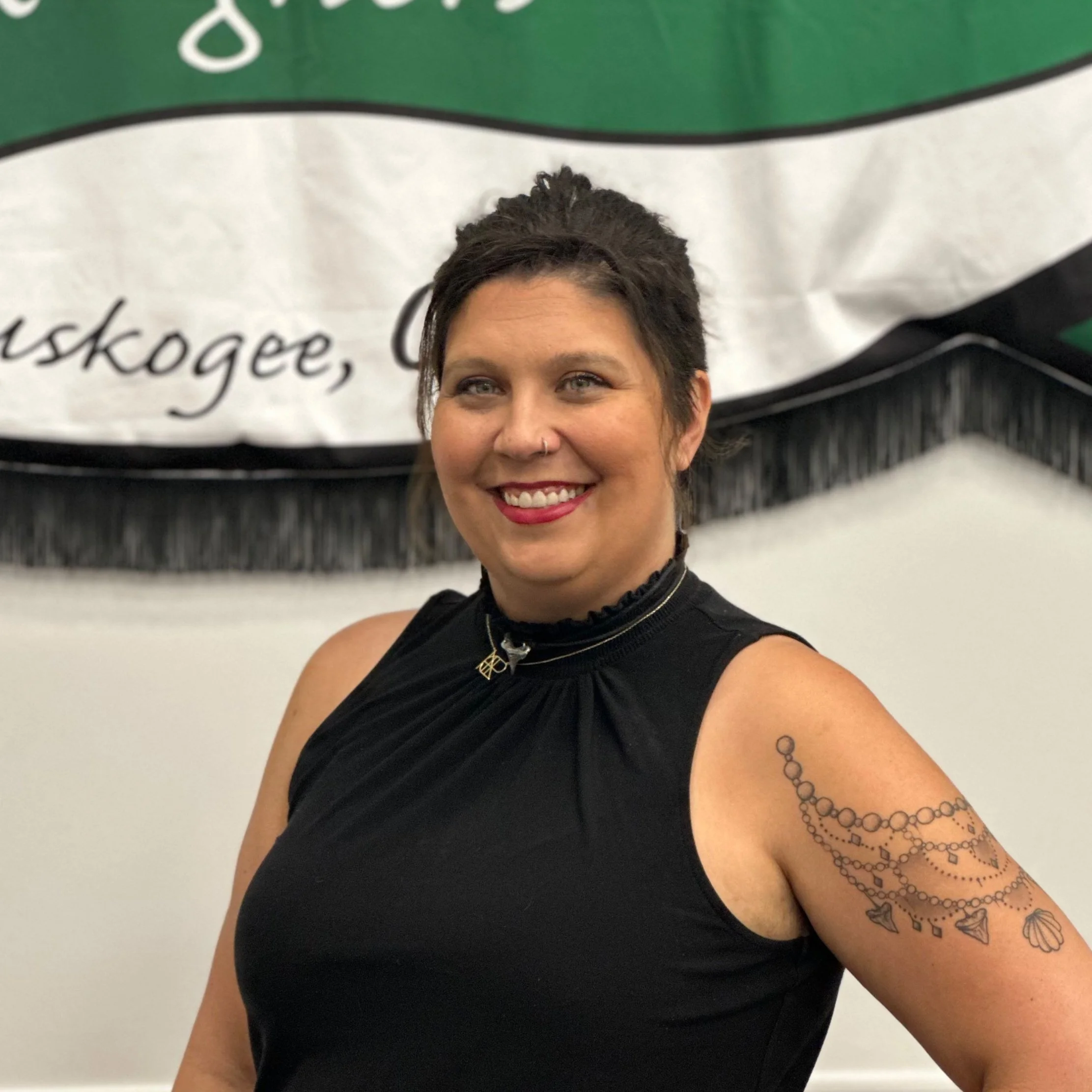 A smiling woman with short dark hair, wearing a sleeveless black top, standing in front of a banner. She has a nose ring, red lipstick, and a tattoo of a shell and geometric shapes on her upper right arm.
