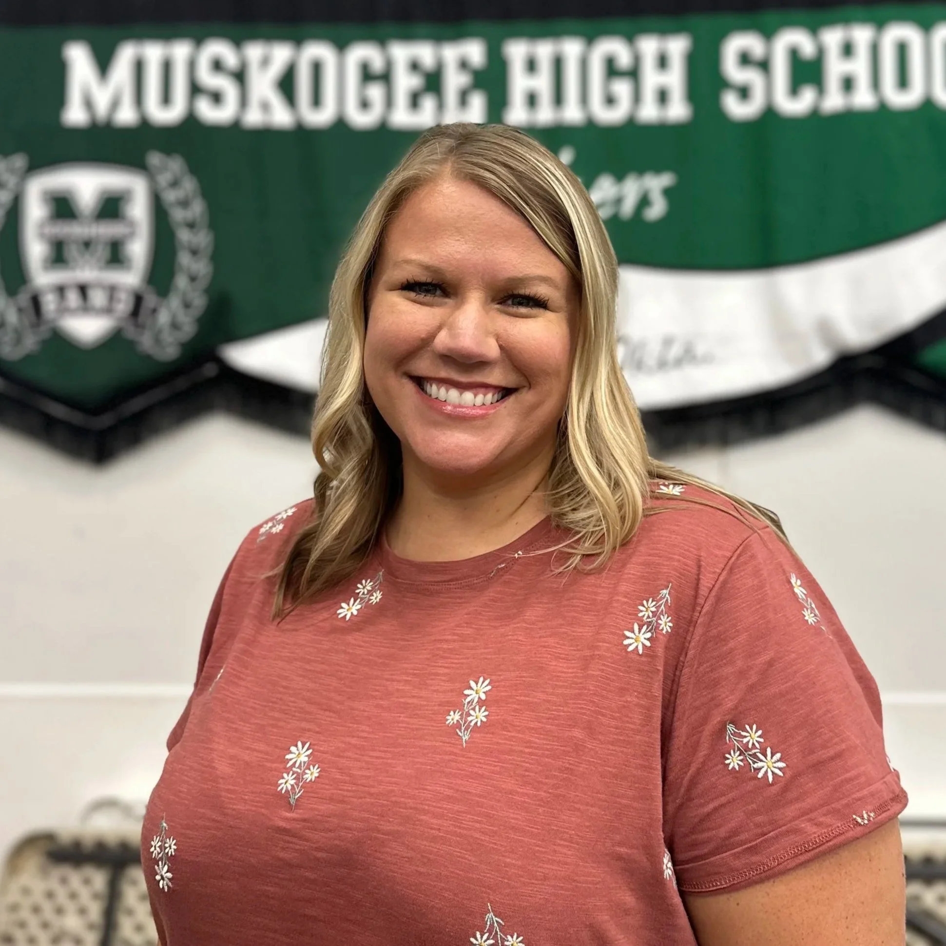 A smiling woman with blonde hair, wearing a red t-shirt with white daisies, standing in front of a green and white Mokogee High School banner.