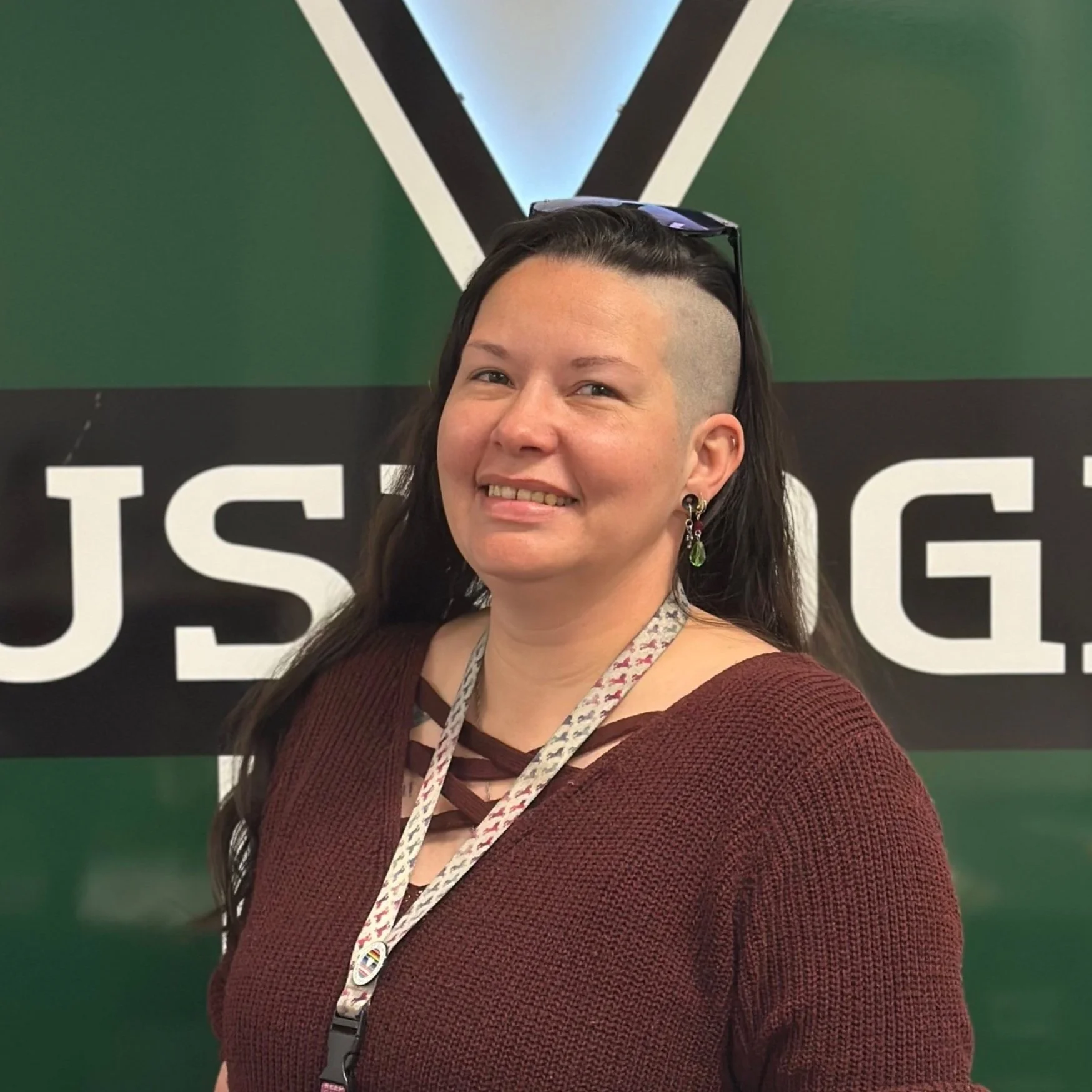 Woman with dark hair styled in a mohawk, wearing a maroon sweater, earrings, and a lanyard, standing in front of a sign with partially visible black and white text.