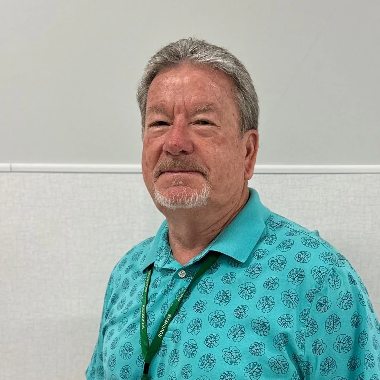 A man with gray hair, a gray goatee, and wearing a turquoise shirt with a leaf pattern, standing in front of a plain light-colored wall.