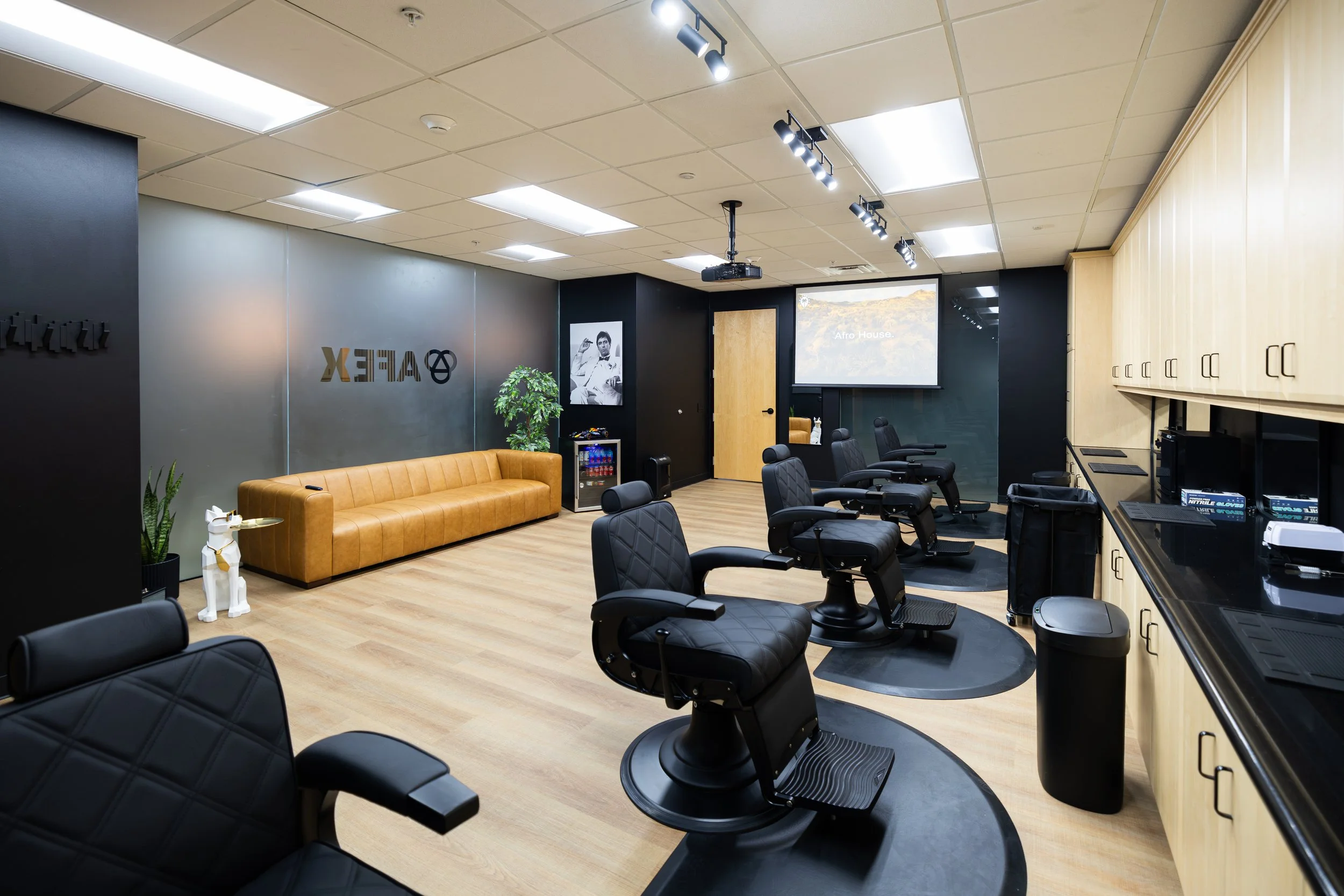 Barber shop with black chairs, a long leather sofa, a TV screen, and hair styling stations.