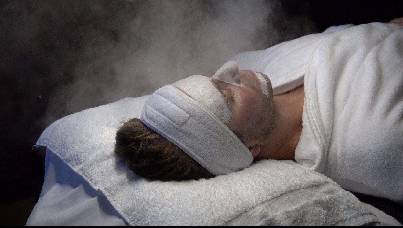 A medical examination room with a white magnifying lamp, three lotion bottles, a rolled black towel, and a black examination table.