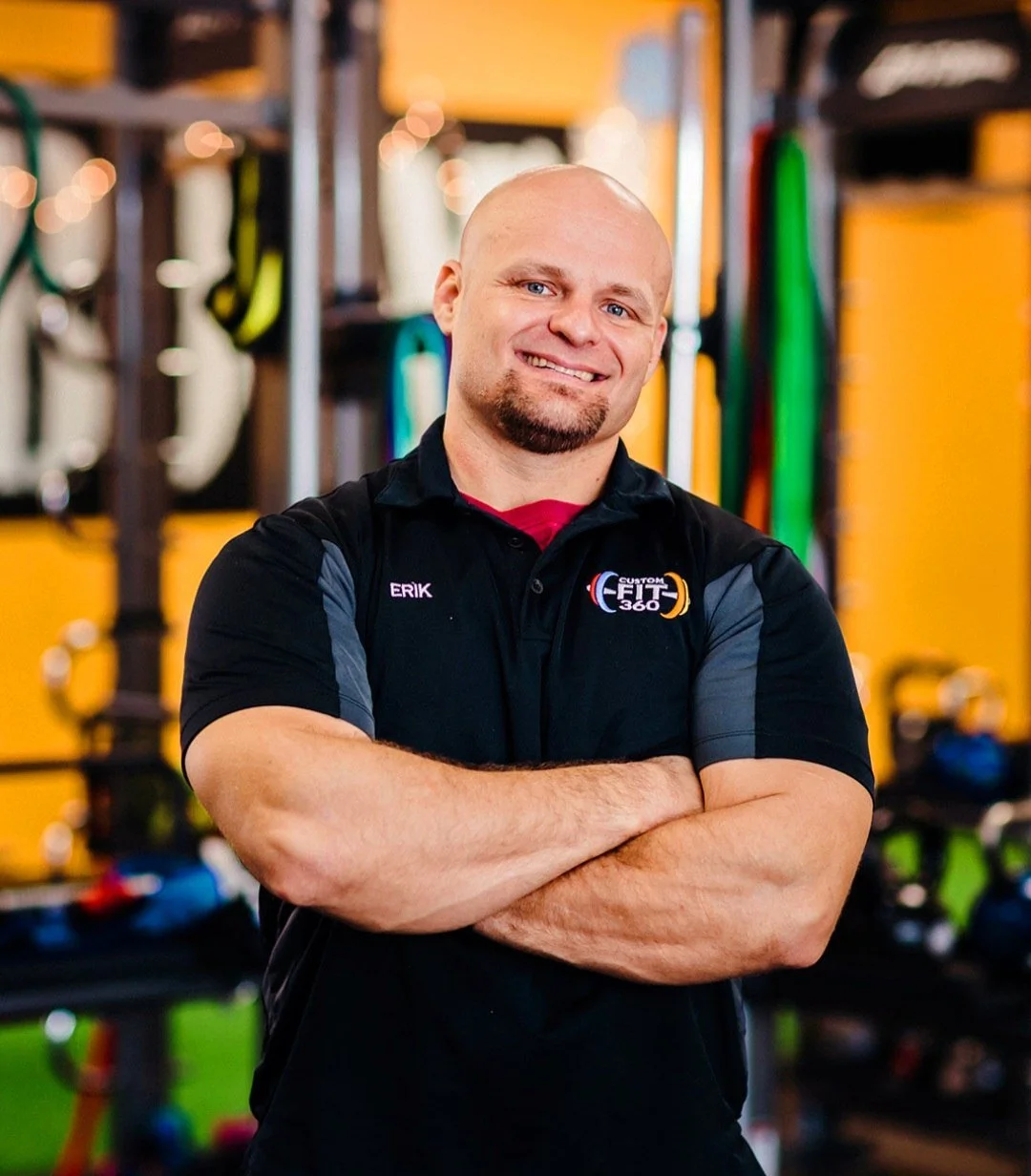 Man wearing a 'Custom Fit 360' shirt in a gym, smiling with arms crossed.