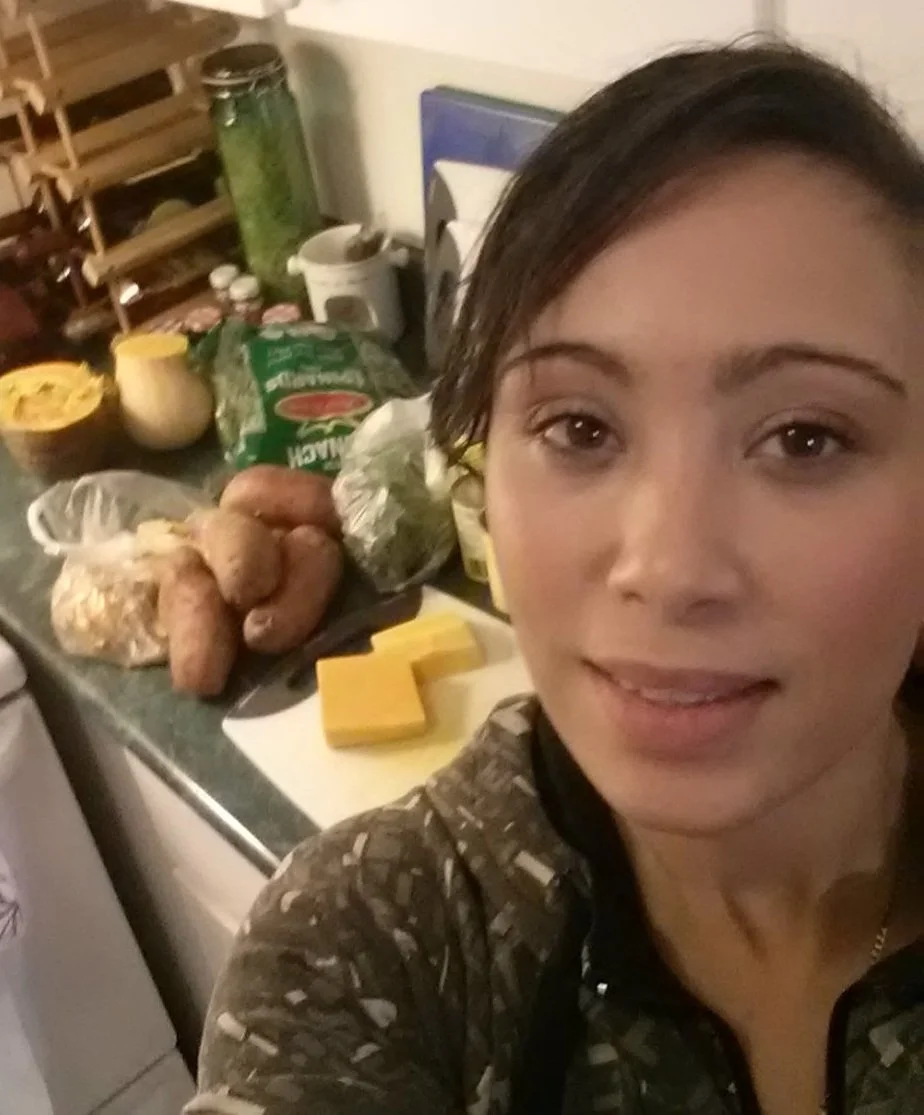 A woman taking a selfie in a kitchen with food ingredients and packaging on the counter behind her, including potatoes, shredded cheese, and condiments.