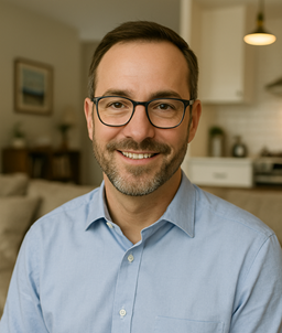 A smiling man with glasses and a beard, wearing a light blue shirt, standing in a cozy living room with a kitchen in the background.